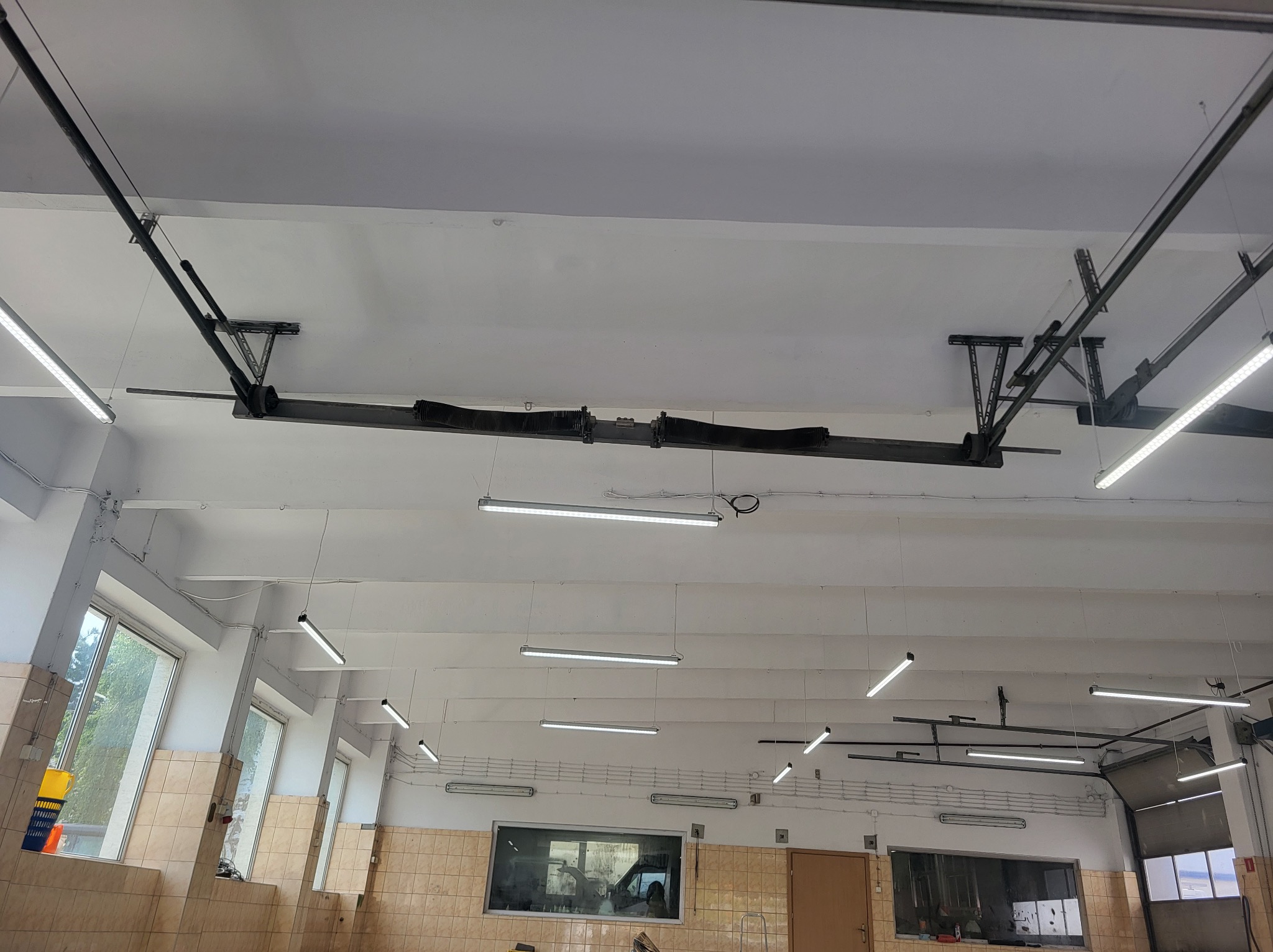 Wnętrze hali warsztatowej z widocznymi elementami konstrukcji bramy segmentowej pod sufitem oraz oświetleniem LED w postaci podłużnych lamp.