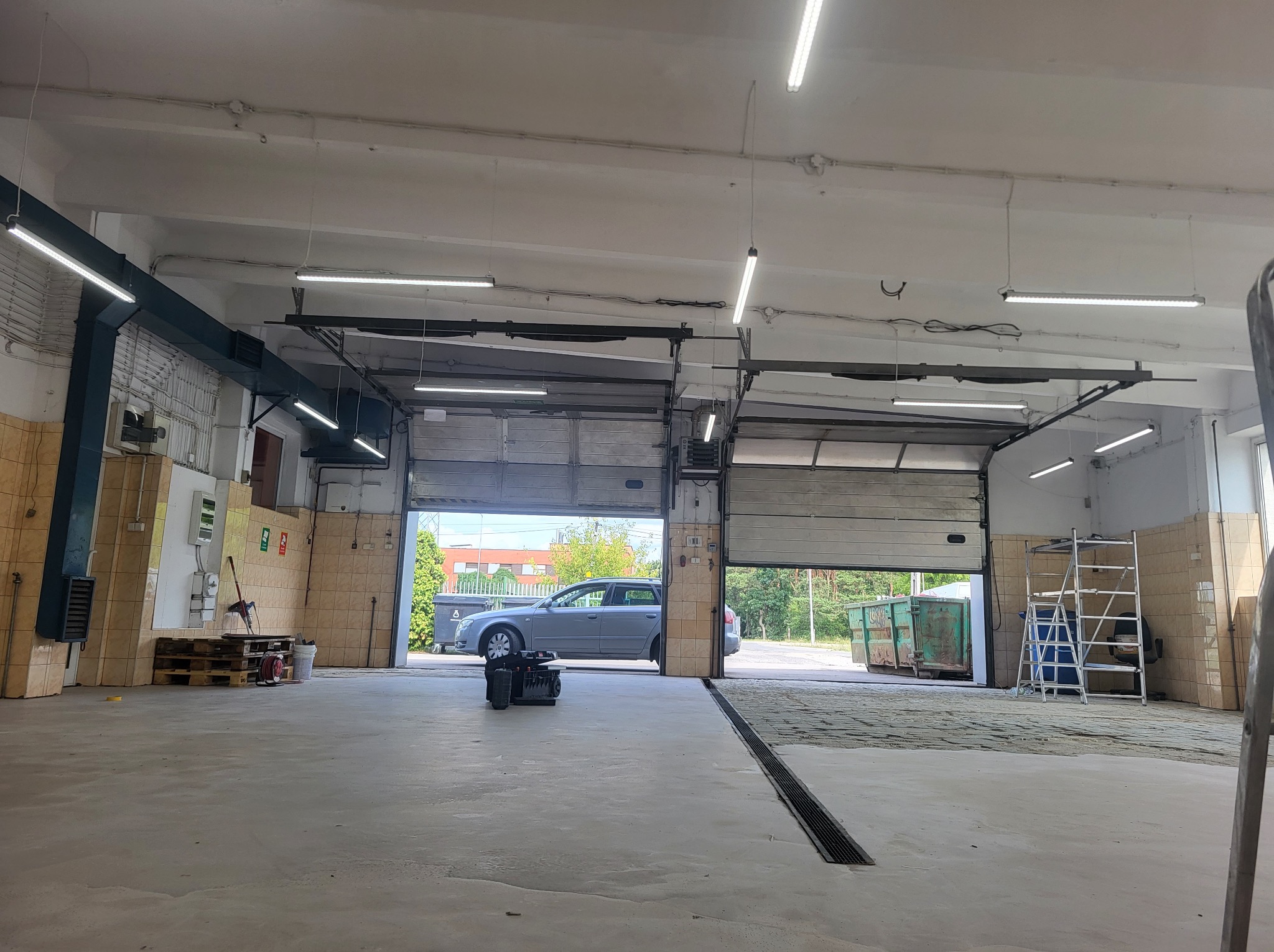 Wnętrze warsztatu z dwoma otwartymi bramami garażowymi, widoczne oświetlenie LED pod sufitem, palety, drabina i narzędzia. Na zewnątrz widoczne samochody i kontenery na śmieci.