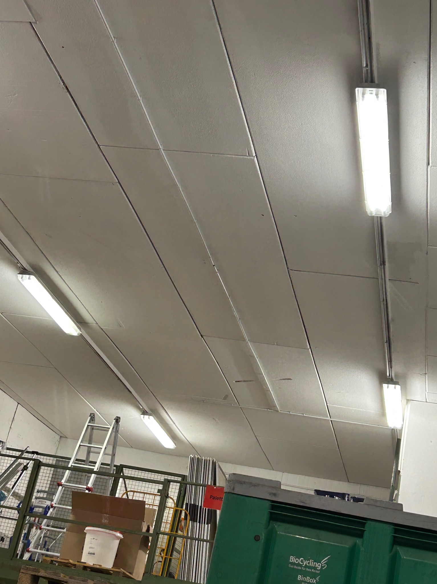 Wnętrze magazynu z widoczną instalacją oświetleniową na suficie z paneli, drabina aluminiowa i pojemnik na odpady BioCycling w dolnym rogu.