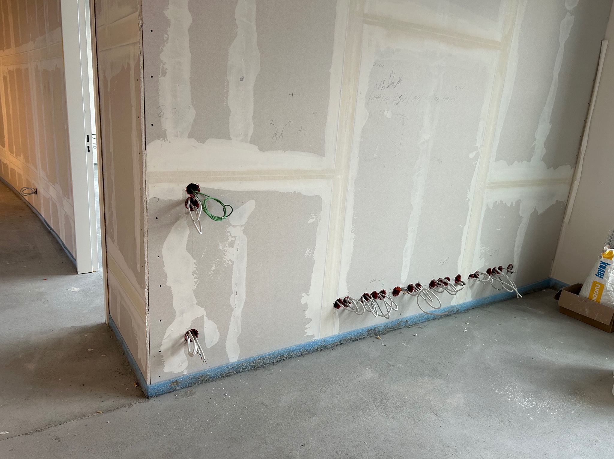 Surowe wnętrze w trakcie remontu: ściany z płyt gipsowo-kartonowych z widocznymi przewodami elektrycznymi wystającymi z otworów, zabezpieczone niebieską taśmą przy podłodze, worek z gipsem w rogu...
