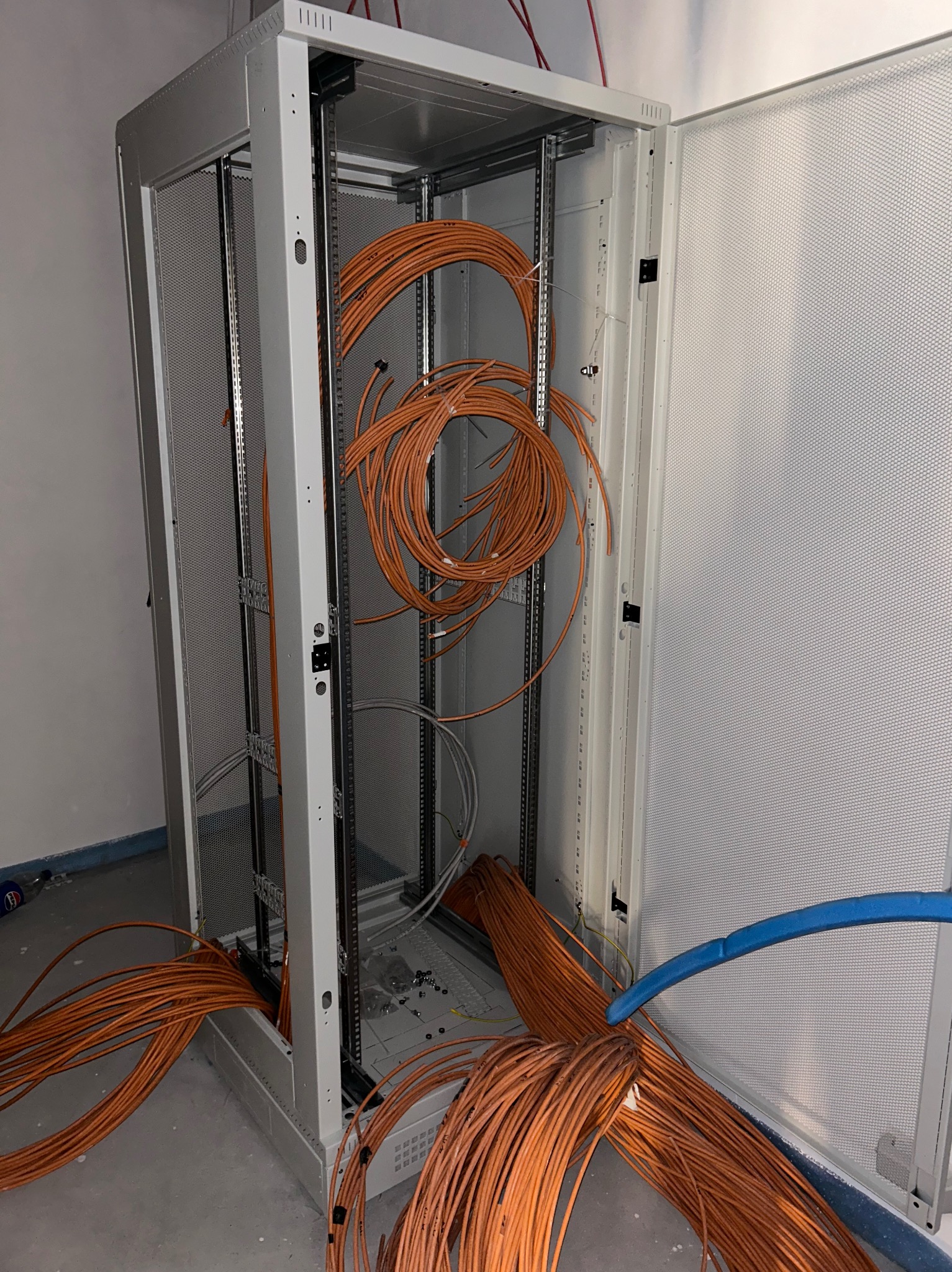 Otwarta szafa serwerowa z pomarańczowymi kablami sieciowymi wewnątrz i na zewnątrz, widoczne metalowe szyny montażowe i perforowane panele boczne.