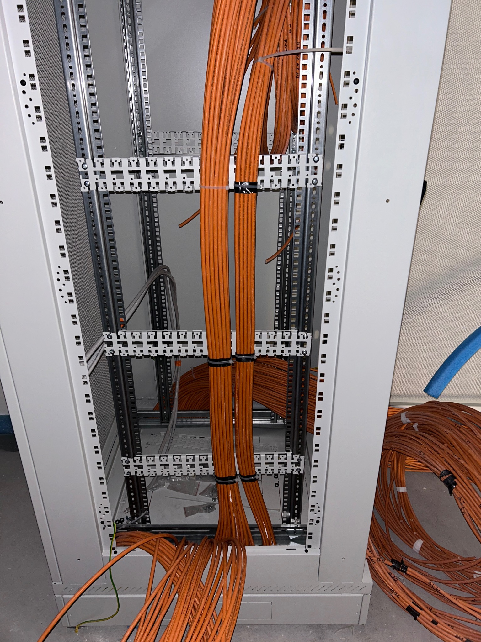Wnętrze szafy serwerowej z uporządkowanymi pomarańczowymi kablami, zabezpieczonymi czarnymi opaskami zaciskowymi, widoczne metalowe elementy montażowe i częściowo zwinięty kabel na podłodze.