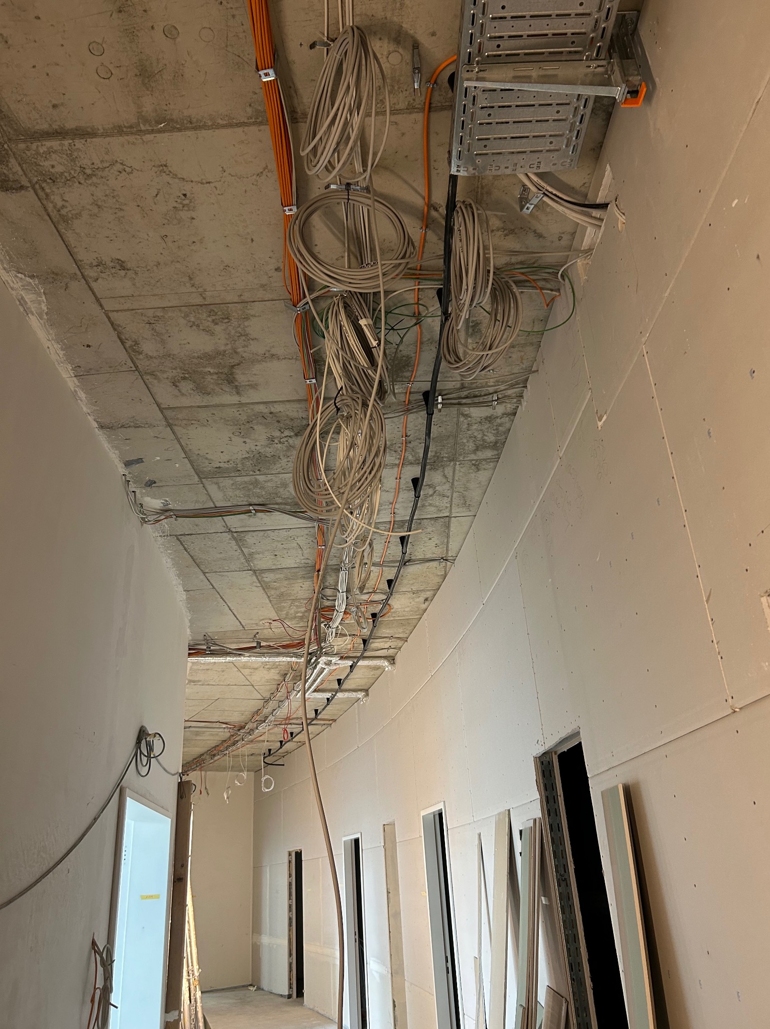 Kable elektryczne i pomarańczowe przewody ułożone pod sufitem w nowym budynku w trakcie instalacji, widoczne koryta kablowe i surowy betonowy sufit.