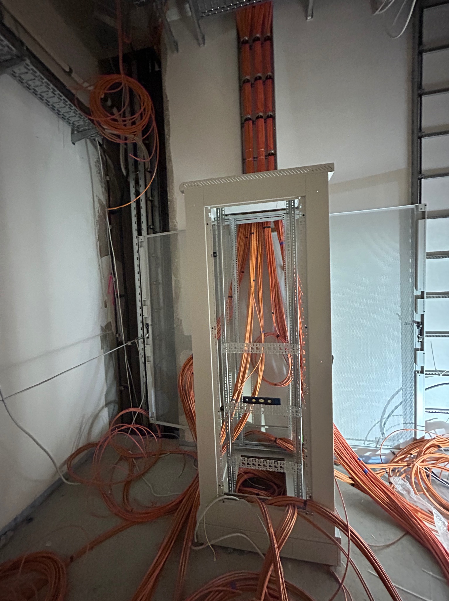 Montaż szafy rack z pomarańczowymi kablami w surowym pomieszczeniu; widoczne kable zwisające z sufitu i leżące na podłodze, częściowo schowane za szafą.