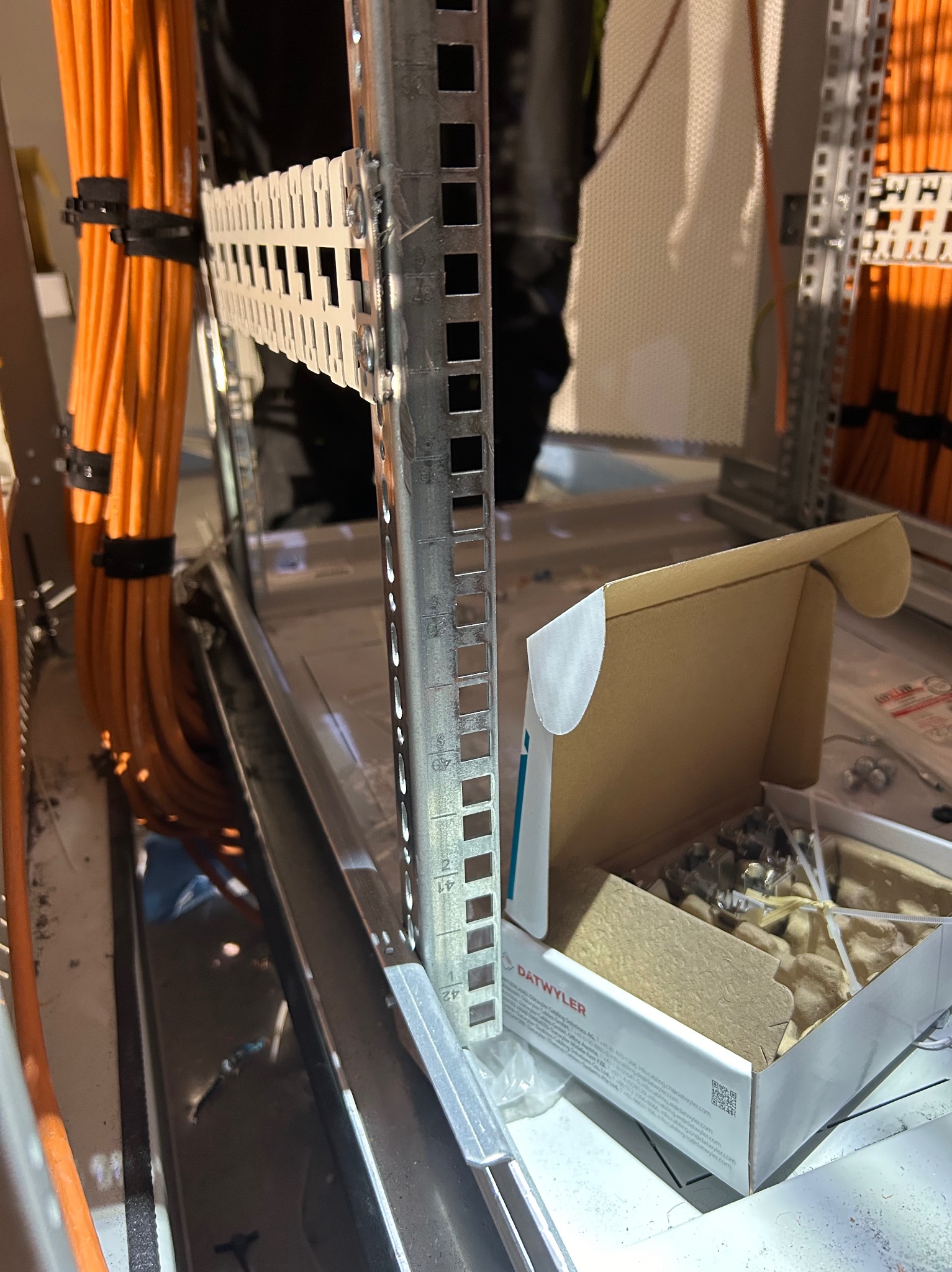 Montaż okablowania strukturalnego w szafie serwerowej, widoczne pomarańczowe kable uporządkowane opaskami, metalowe szyny montażowe z perforacją i pudełko z elementami mocującymi marki Datwyler.