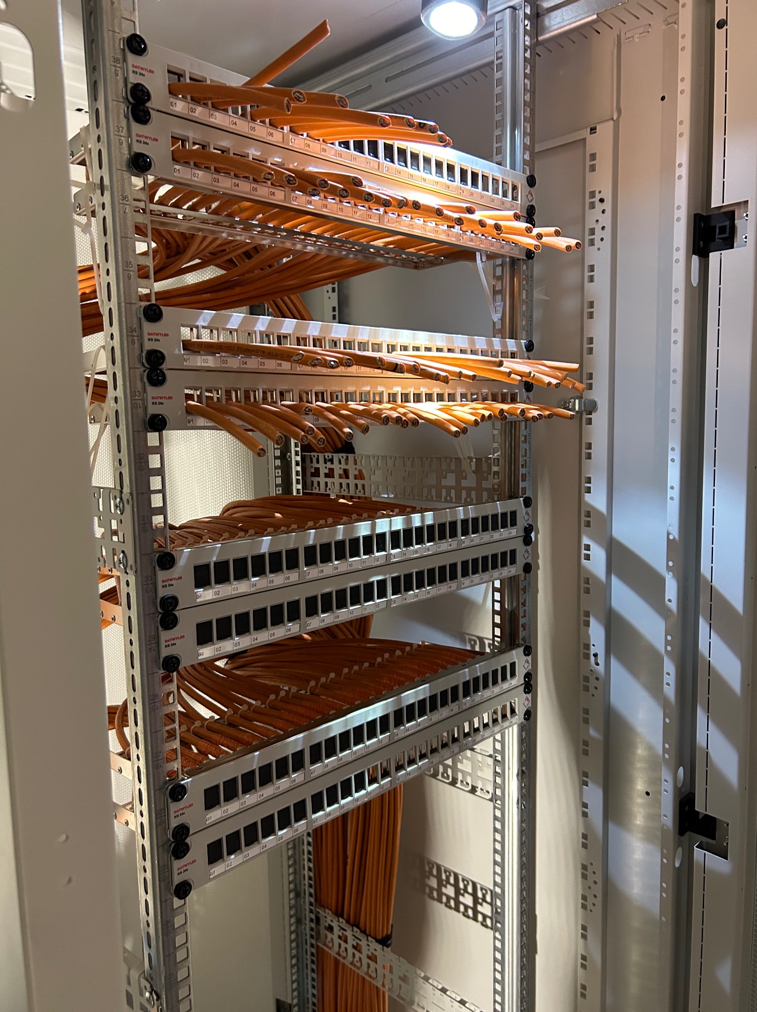 Pomarańczowe kable sieciowe uporządkowane w szafie rack, widoczne panele krosowe z numerowanymi portami, oświetlenie górne.