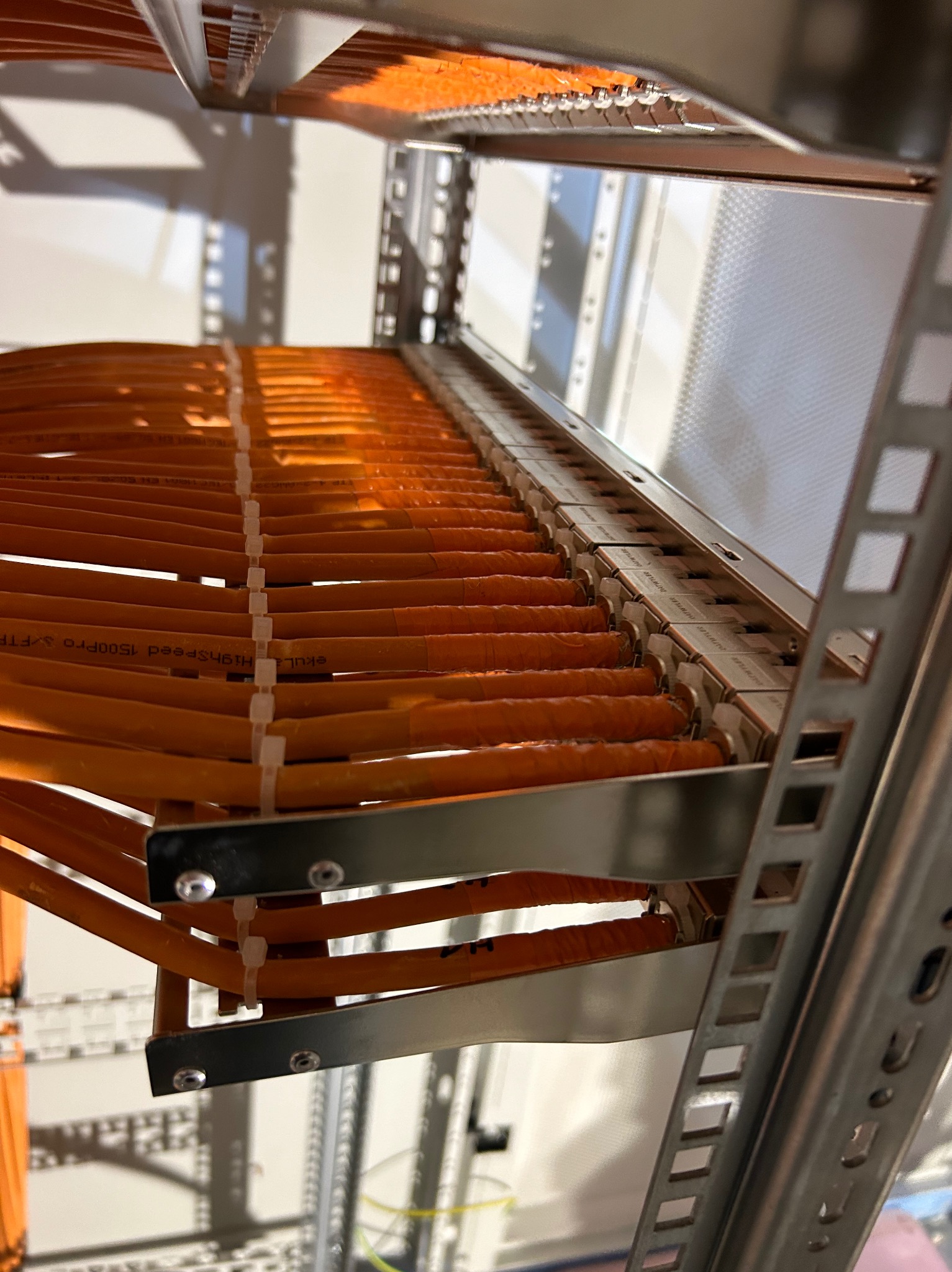 Rząd pomarańczowych kabli sieciowych Highspeed 1500PRO w metalowej szafie rack, spiętych białymi opaskami zaciskowymi, widok z dołu pod kątem.