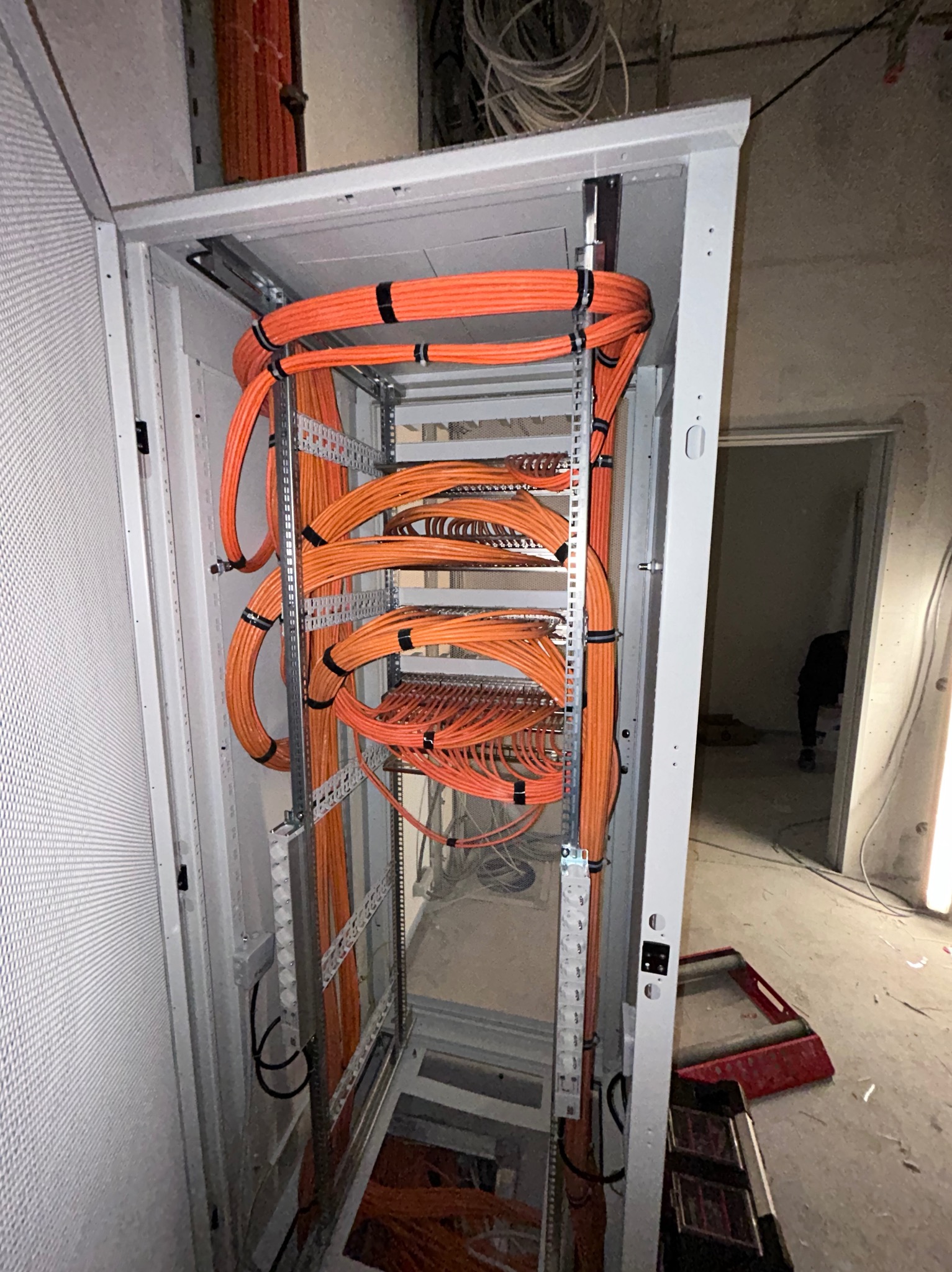 Pomarańczowe kable elektryczne uporządkowane w szafie rack, zabezpieczone czarnymi opaskami zaciskowymi, widoczne elementy konstrukcyjne szafy i fragment surowego wnętrza budynku w tle.