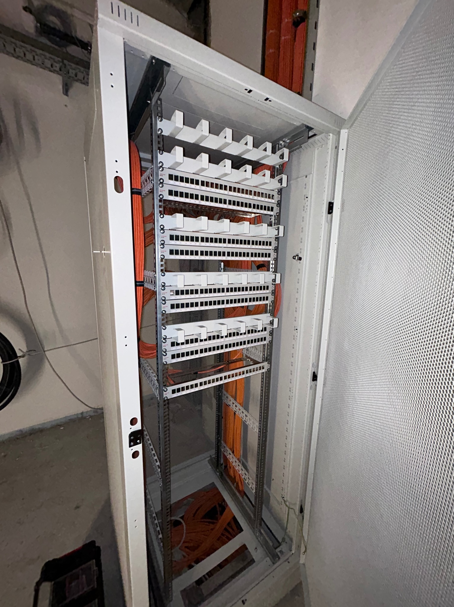 Otwarta szafa rack z uporządkowanymi pomarańczowymi kablami, widoczne białe uchwyty kablowe i metalowa konstrukcja, w tle surowy beton.
