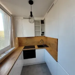 Nowoczesna kuchnia z białymi, błyszczącymi szafkami, drewnianym blatem i panelem ściennym, czarnym zlewem i płytą indukcyjną, oświetlona dekoracyjną lampą wiszącą.