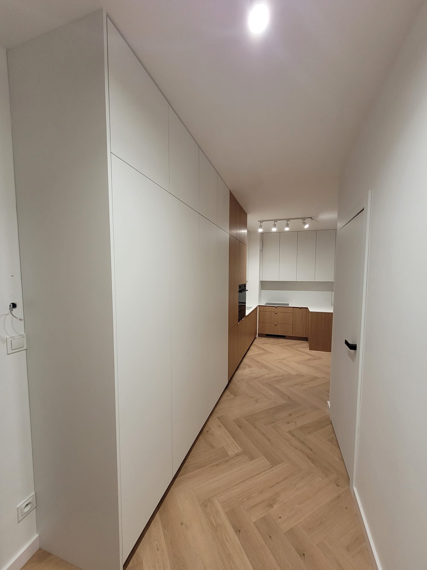 Biała zabudowa szafy w korytarzu przechodząca w zabudowę kuchenną z drewnianymi akcentami. Parkiet ułożony w jodełkę. Minimalistyczny design.