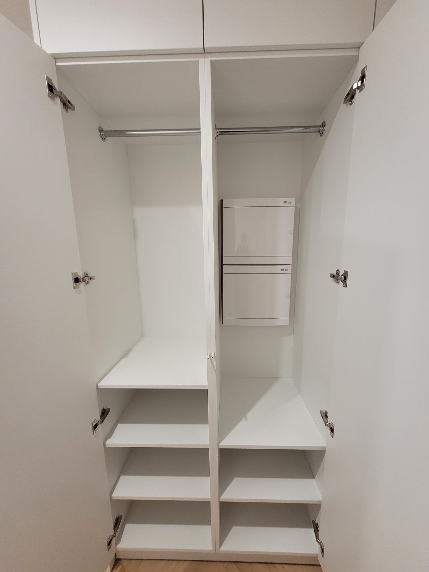 Wnętrze białej szafy wnękowej z półkami i drążkiem na ubrania, widoczna skrzynka elektryczna. Minimalistyczny design, otwarte drzwi ukazują jasne wnętrze.