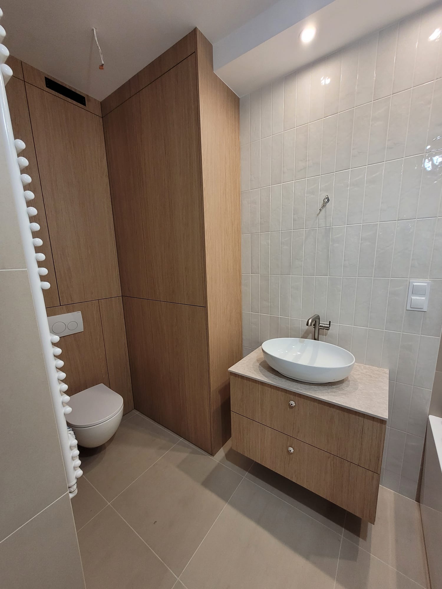 Nowoczesna łazienka z drewnianą zabudową meblową, umywalką nablatową i grzejnikiem drabinkowym. Jasne płytki na ścianach i podłodze. Minimalistyczny design.