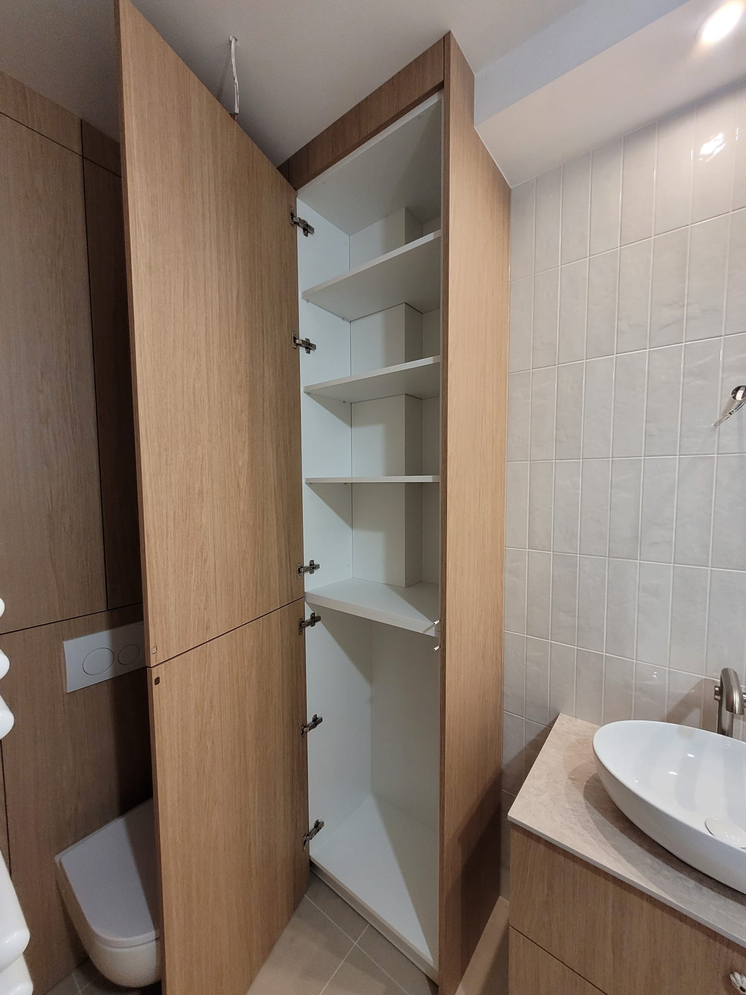 Wysoka, drewniana szafa łazienkowa z otwartymi drzwiami, ukazująca białe półki. Obok umywalka i toaleta. Jasne płytki na ścianie. Minimalistyczny design.