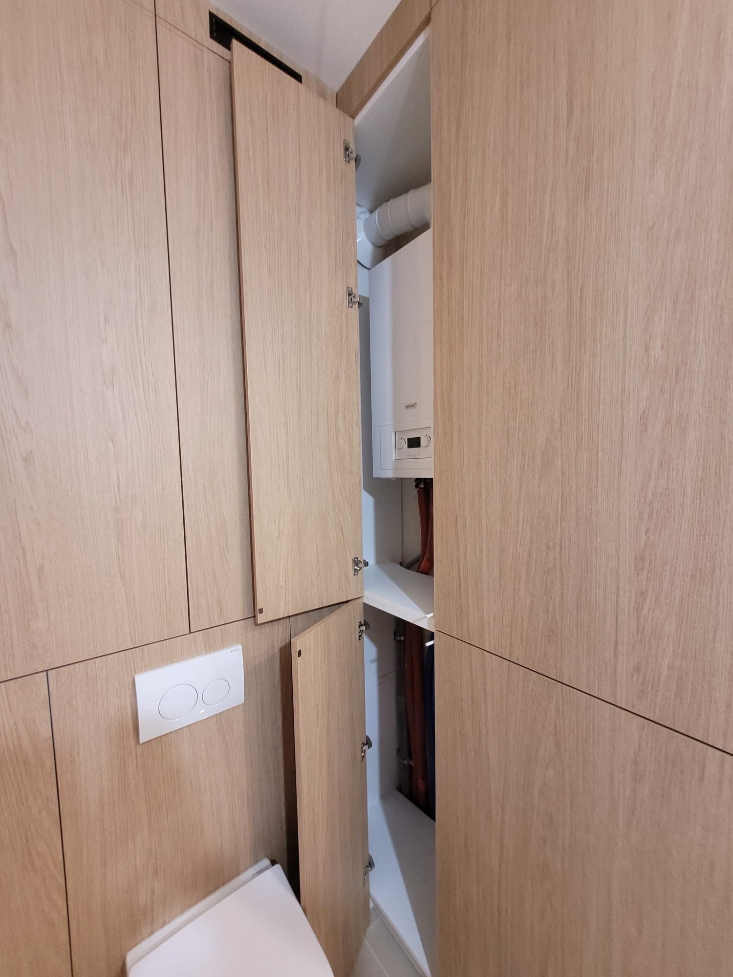 Jasne, drewniane szafki na wymiar z ukrytym piecem gazowym i toaletą. Minimalistyczny design, otwarte drzwiczki ukazują wnętrze szafki.