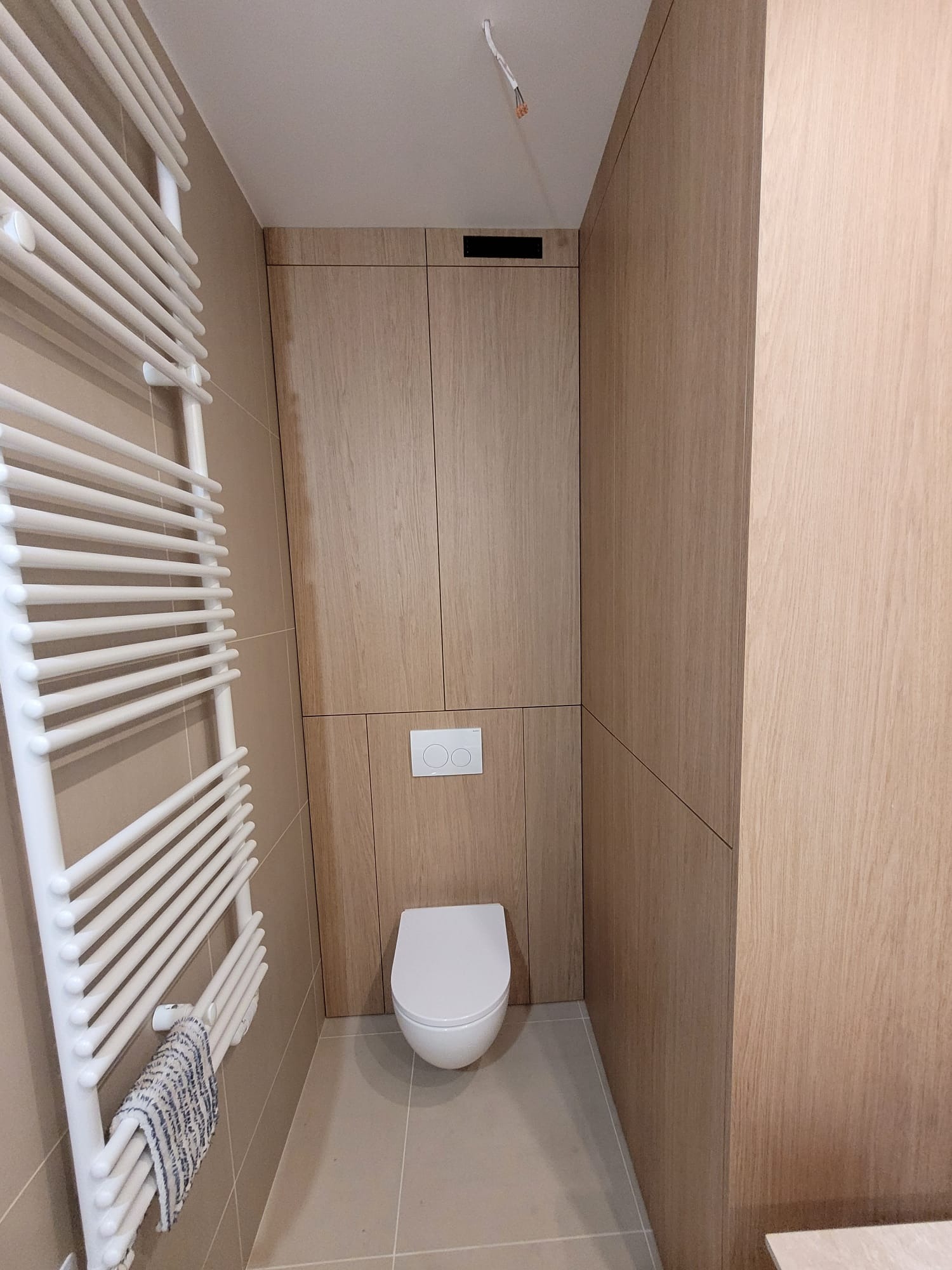 Nowoczesna toaleta z zabudową z jasnego drewna, wiszącą muszlą i grzejnikiem drabinkowym z ręcznikiem. Minimalistyczny design, ciepłe odcienie, subtelne oświetlenie.