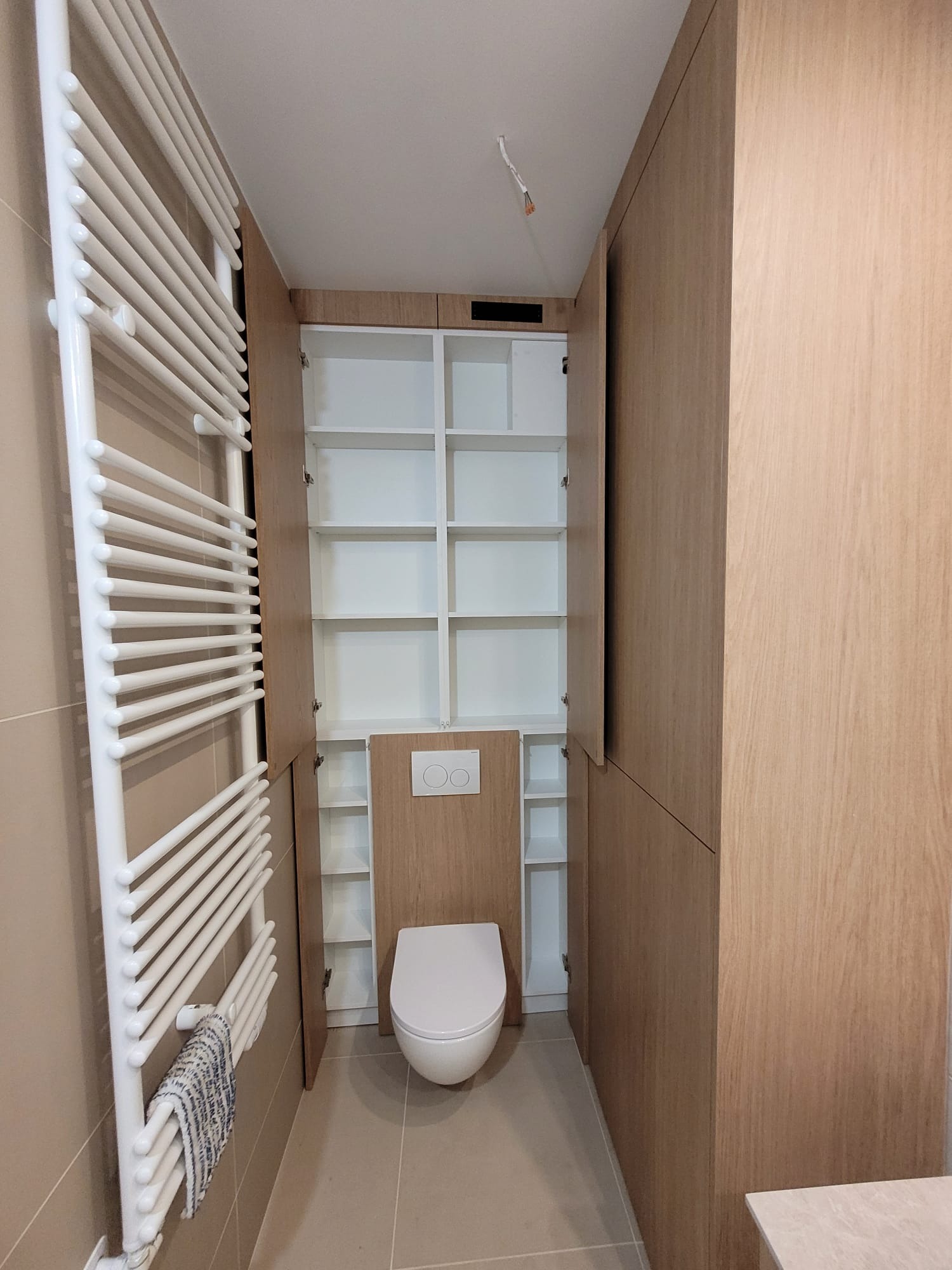 Nowoczesna toaleta z zabudową meblową z drewna w odcieniach beżu, z otwartymi szafkami i grzejnikiem drabinkowym. Minimalistyczny design, jasne wnętrze.