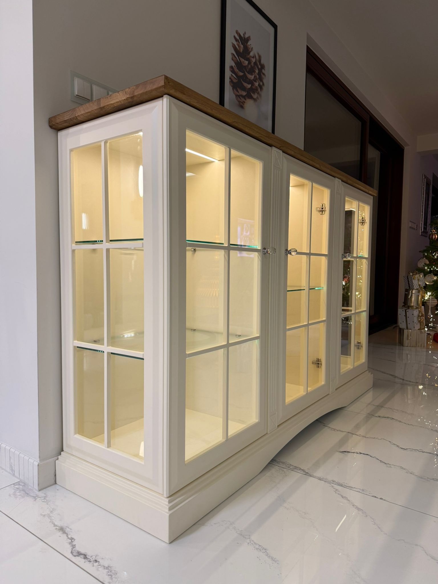 Biała witryna z drewnianym blatem, przeszklonymi drzwiami i oświetleniem wewnętrznym, ustawiona na błyszczącej podłodze. W tle widoczna dekoracja świąteczna.