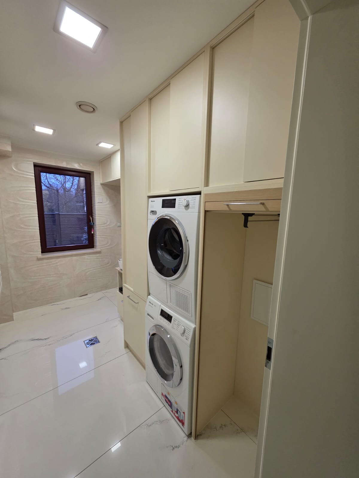 Jasne szafy w zabudowie z pralką i suszarką w łazience. Widoczne białe płytki podłogowe i okno z widokiem na zewnątrz. Minimalistyczny design.