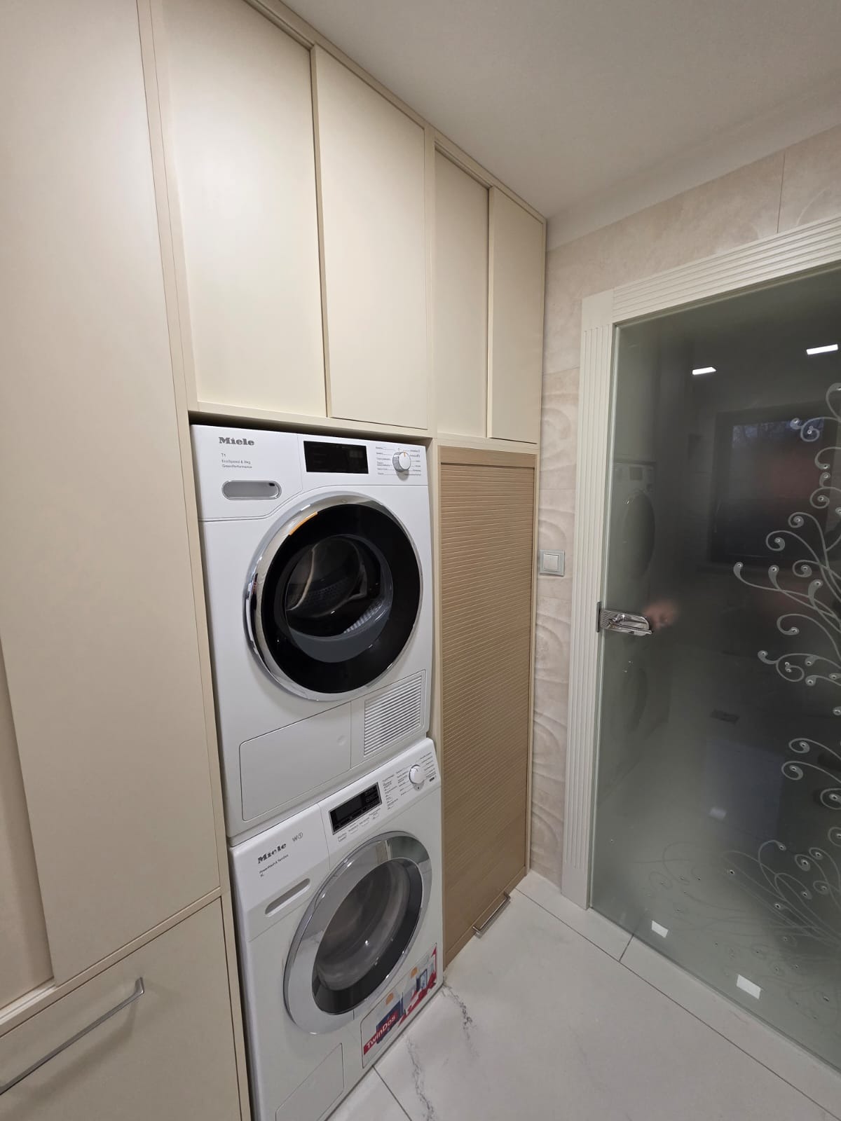 Biała pralka i suszarka Miele w zabudowie meblowej z beżowymi frontami, obok drzwi z ornamentem. Ujęcie z dołu, widoczne płytki podłogowe.