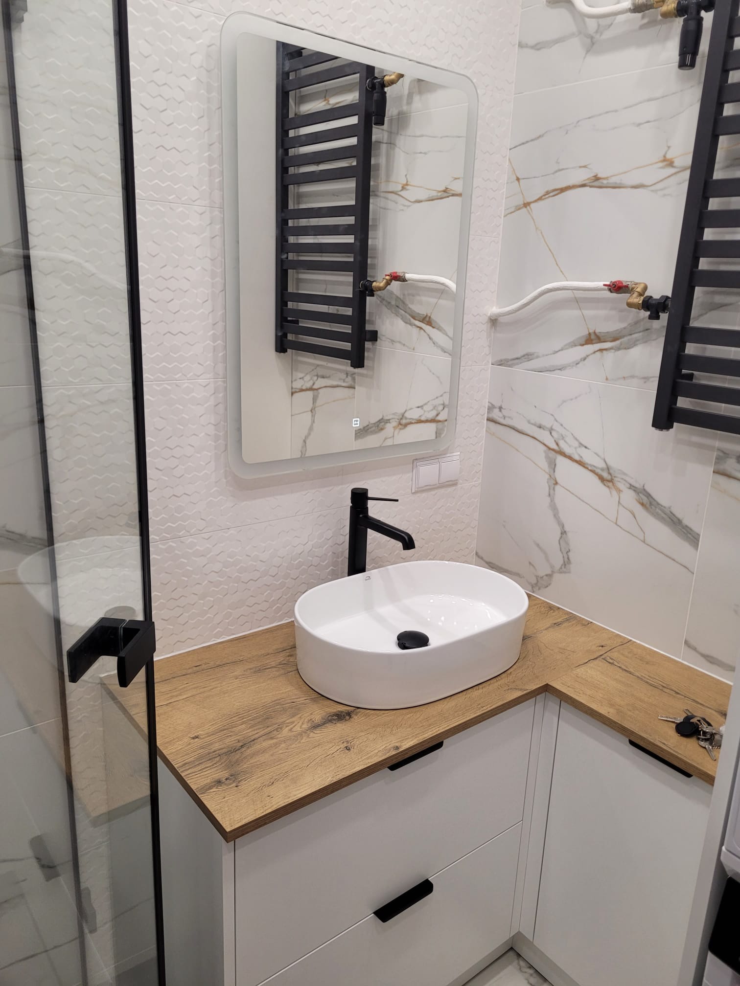 Nowoczesna łazienka z białą umywalką nablatową na blacie drewnopodobnym, czarną armaturą i grzejnikiem drabinkowym. Białe płytki z delikatnym wzorem i marmurowym akcentem.