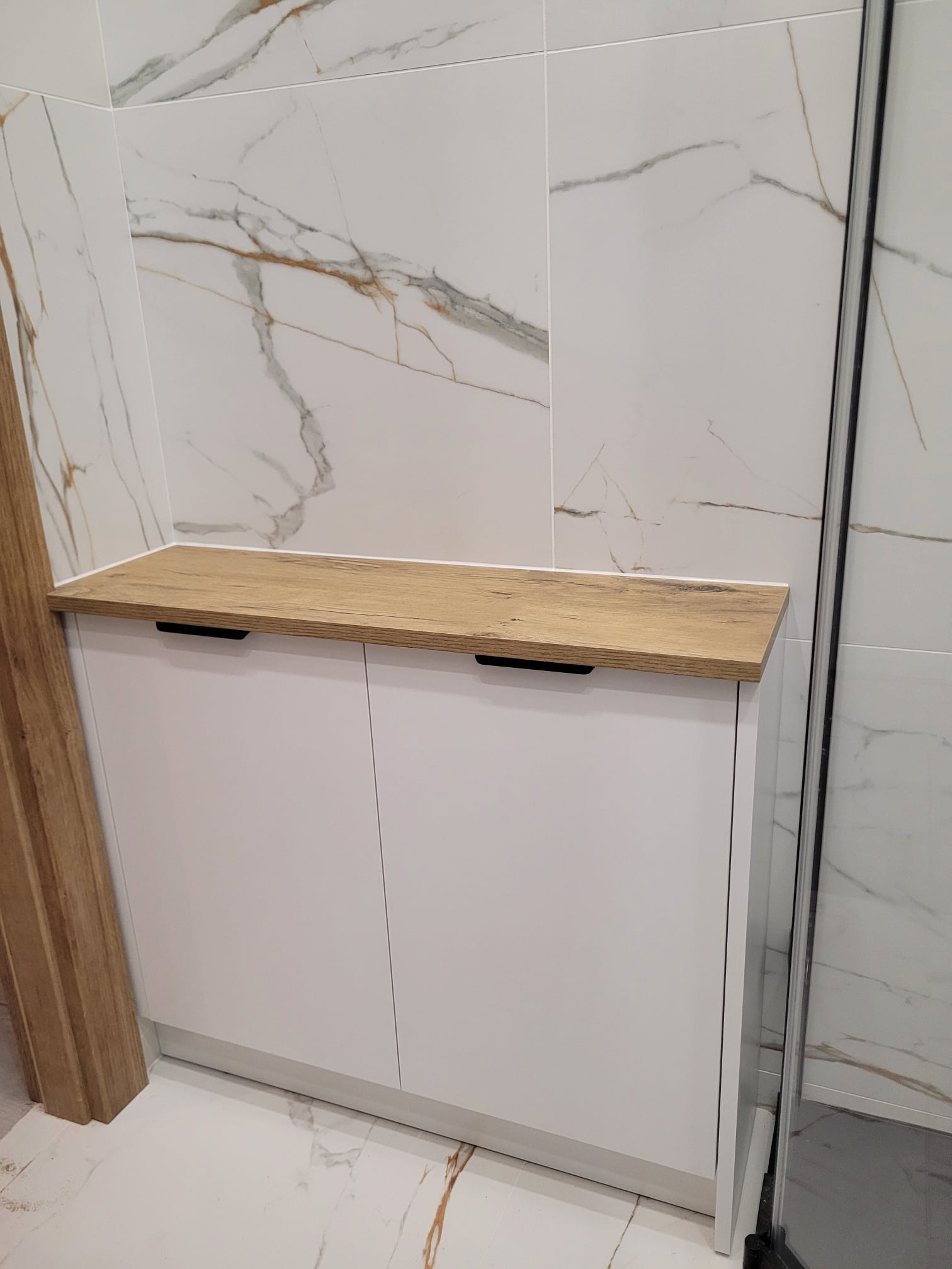 Biała szafka łazienkowa z blatem drewnopodobnym na tle marmurowych płytek. Minimalistyczny design, czarne uchwyty, widoczny fragment kabiny prysznicowej.