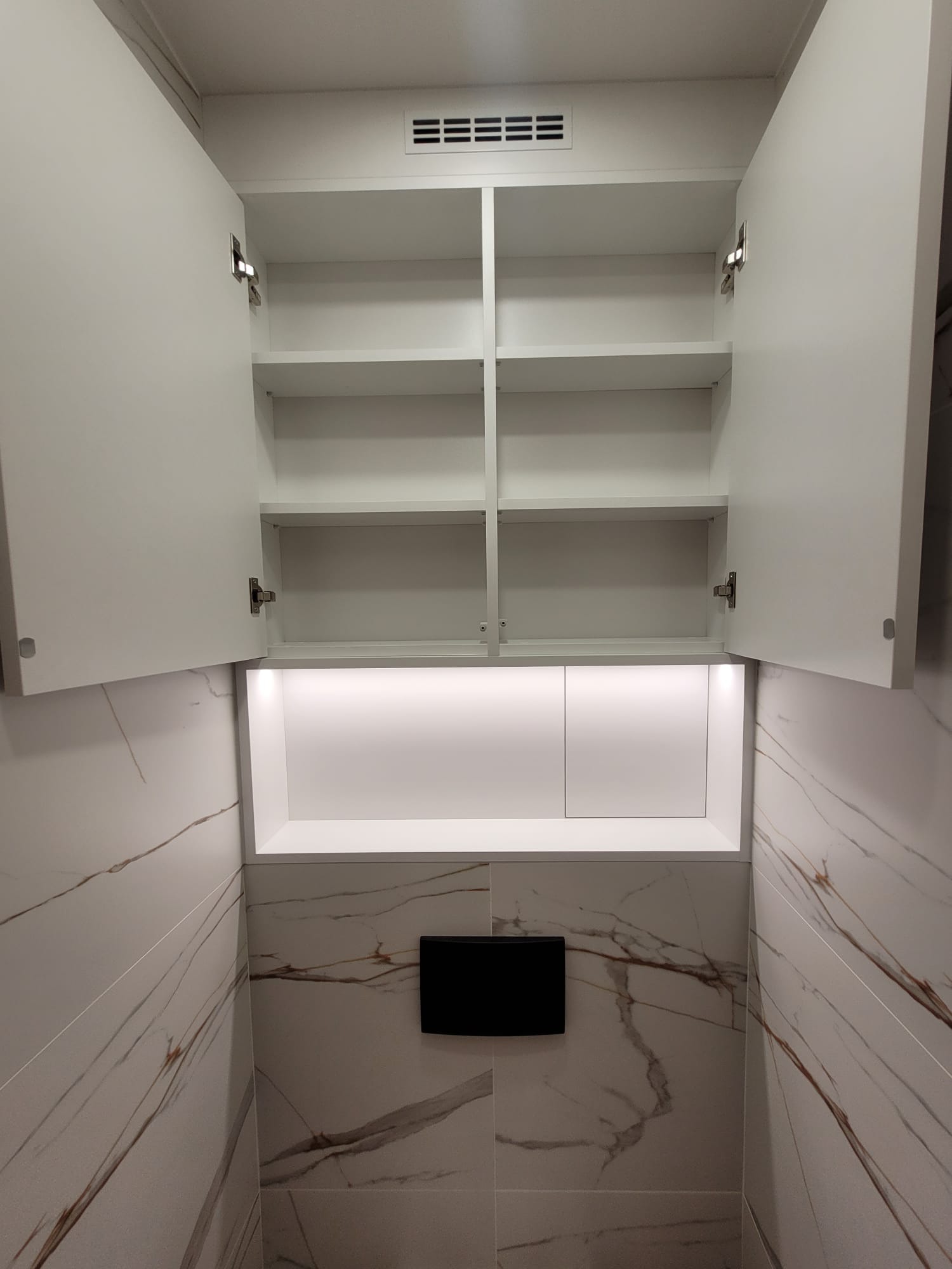 Biała szafka wnękowa z otwartymi drzwiczkami, oświetleniem LED i półkami, zamontowana nad toaletą na ścianie wyłożonej płytkami imitującymi marmur.