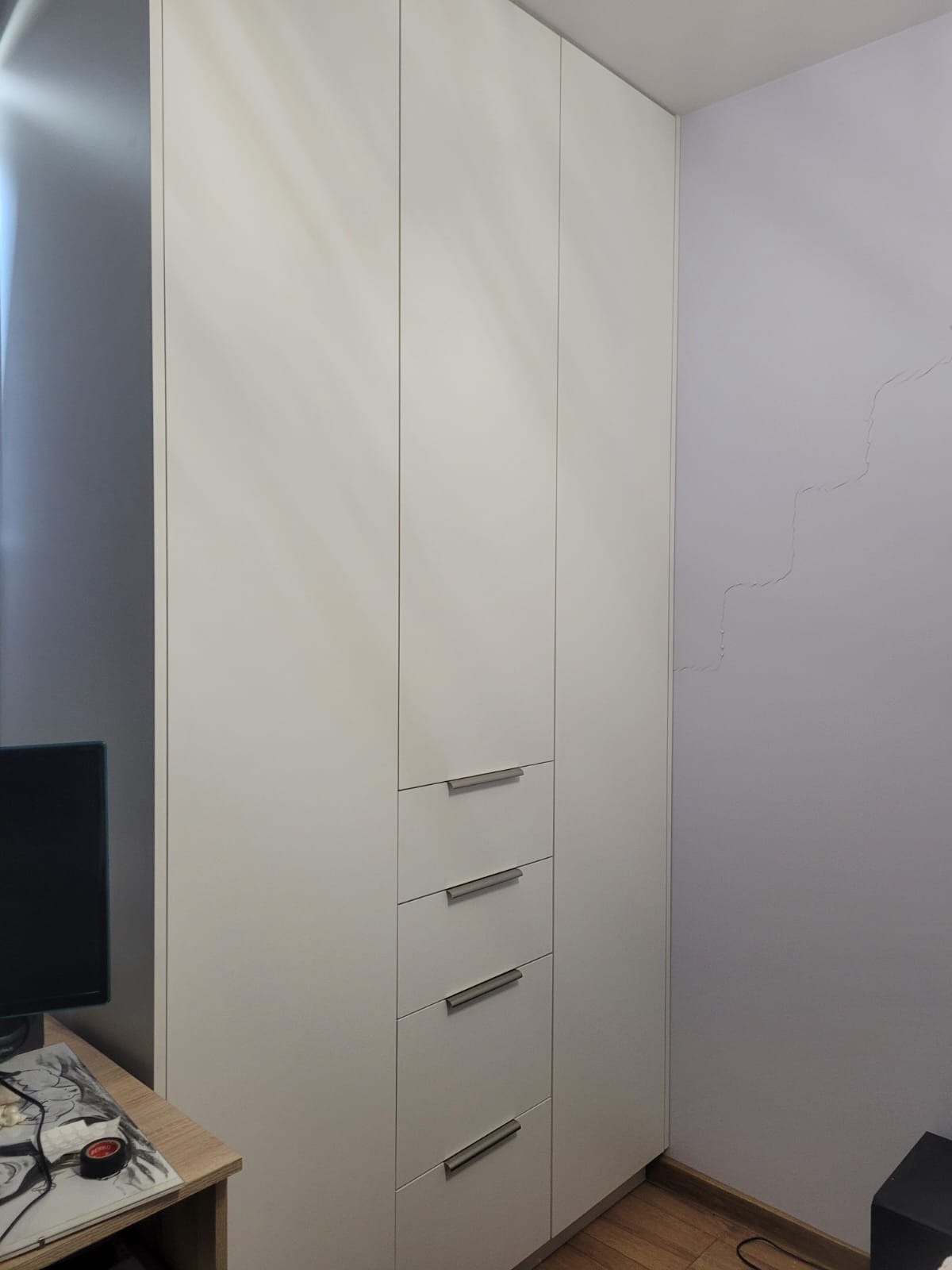 Biała szafa wnękowa z trzema szufladami i czterema drzwiami, z minimalistycznymi uchwytami, ustawiona w rogu pokoju z jasnymi ścianami i drewnianą podłogą.