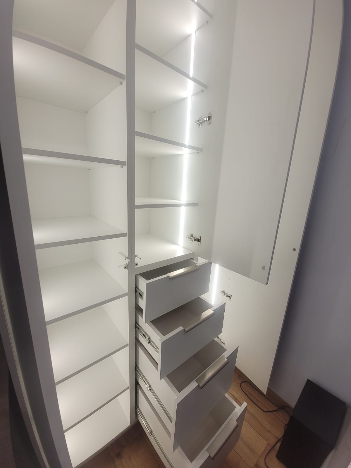 Biała szafa wnękowa z oświetleniem LED, otwartymi szufladami i półkami. Widoczne okucia i detale wykończenia. Ujęcie z dołu, ukazujące wnętrze szafy.