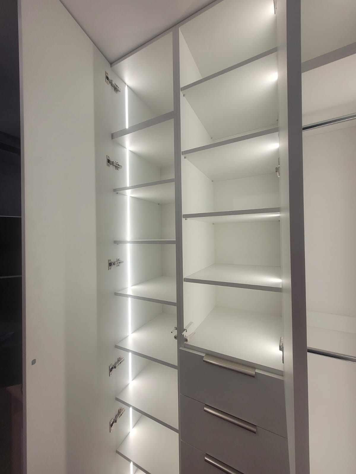 Biała szafa wnękowa z oświetleniem LED, otwarte półki i szuflady z metalowymi uchwytami. Widoczne zawiasy i elementy montażowe. Minimalistyczny design.