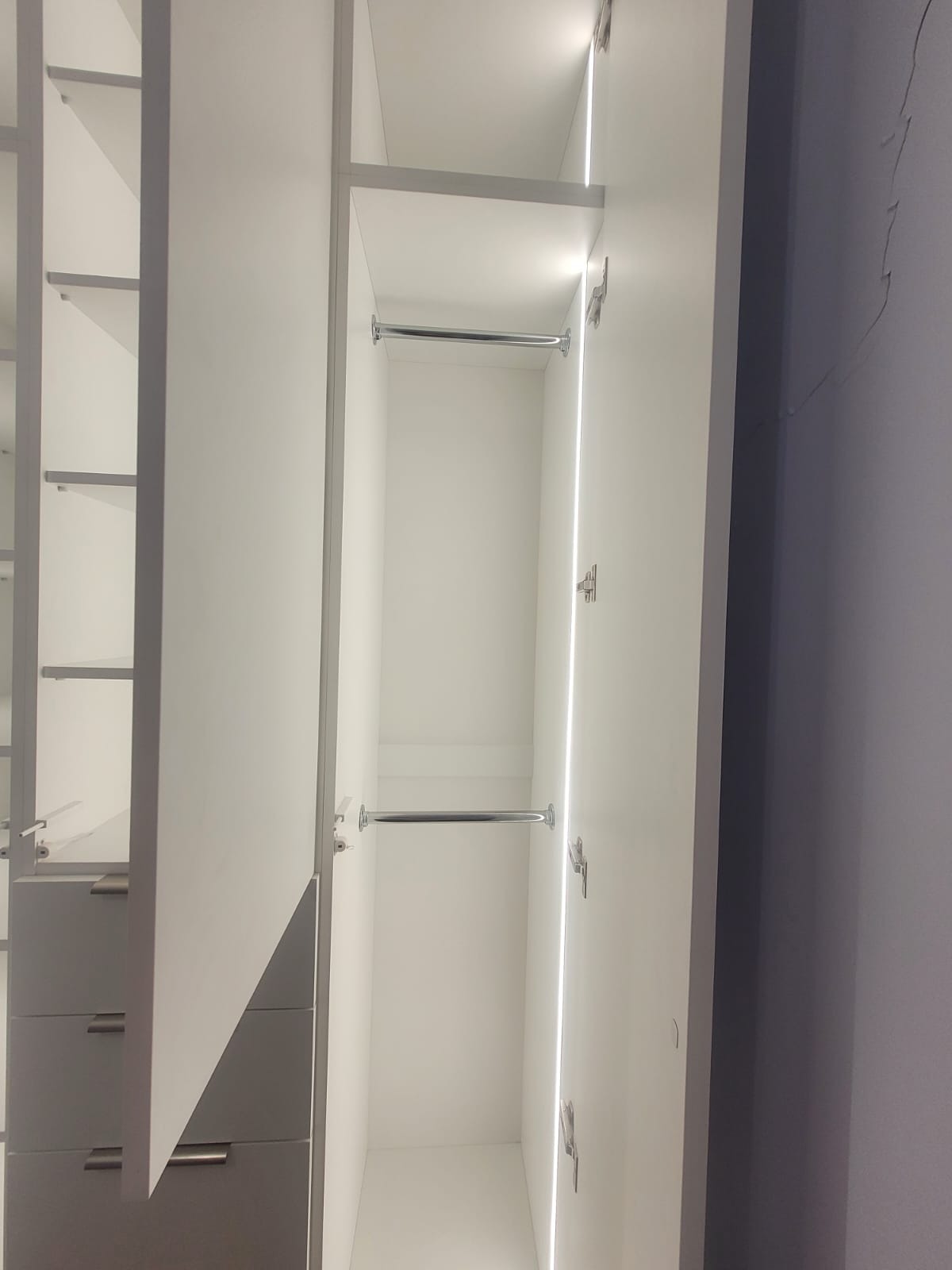 Wnętrze szafy wnękowej z oświetleniem LED, widoczne półki i drążki na ubrania. Minimalistyczny design w odcieniach bieli i szarości. Częściowo otwarte drzwi.