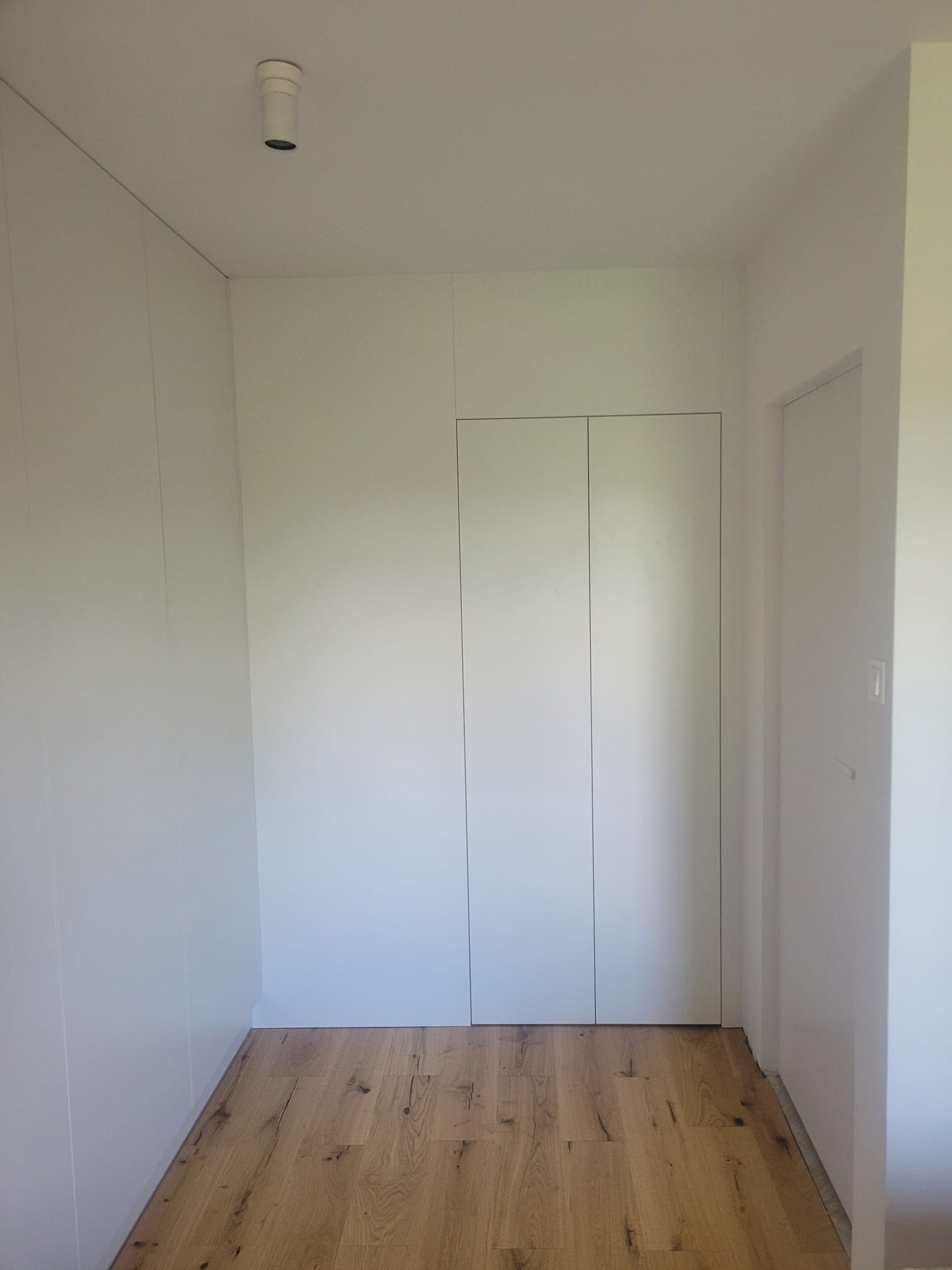 Minimalistyczna, biała szafa wnękowa z ukrytymi drzwiami, wkomponowana w ścianę, na tle drewnianej podłogi i białych ścian. Prosty design, brak klamek.