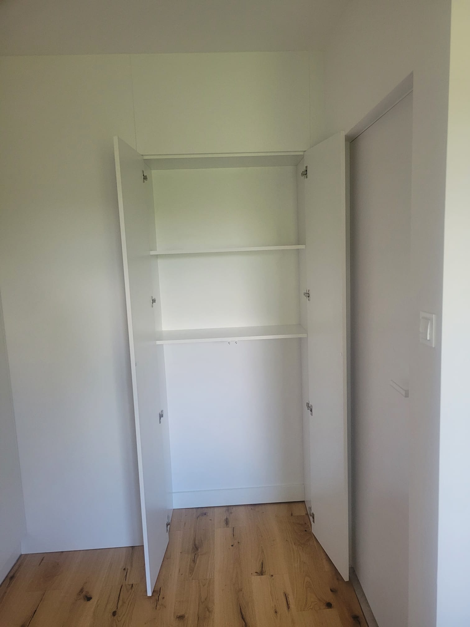 Wnętrze białej szafy wnękowej z półkami, otwarte drzwi ukazują drewnianą podłogę. Minimalistyczny design, jasne wnętrze, widoczne zawiasy.