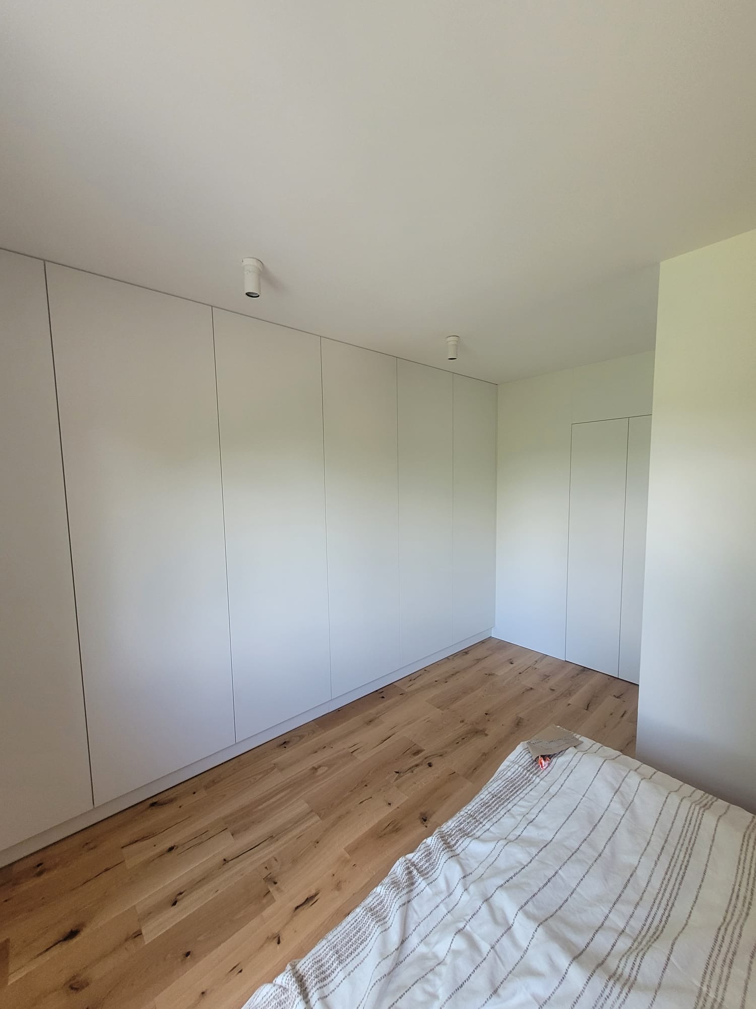 Minimalistyczna sypialnia z zabudowaną szafą w kolorze białym, drewnianą podłogą i fragmentem łóżka z pościelą w paski. Oświetlenie punktowe na suficie.