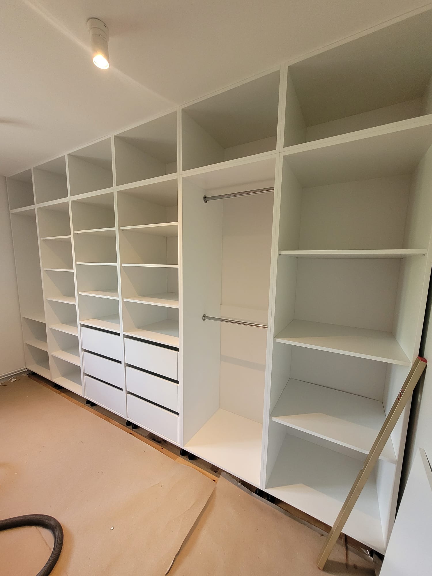 Biała, modułowa szafa wnękowa z szufladami i półkami, widok z dołu, w trakcie montażu, z widocznymi elementami wykończeniowymi na podłodze.