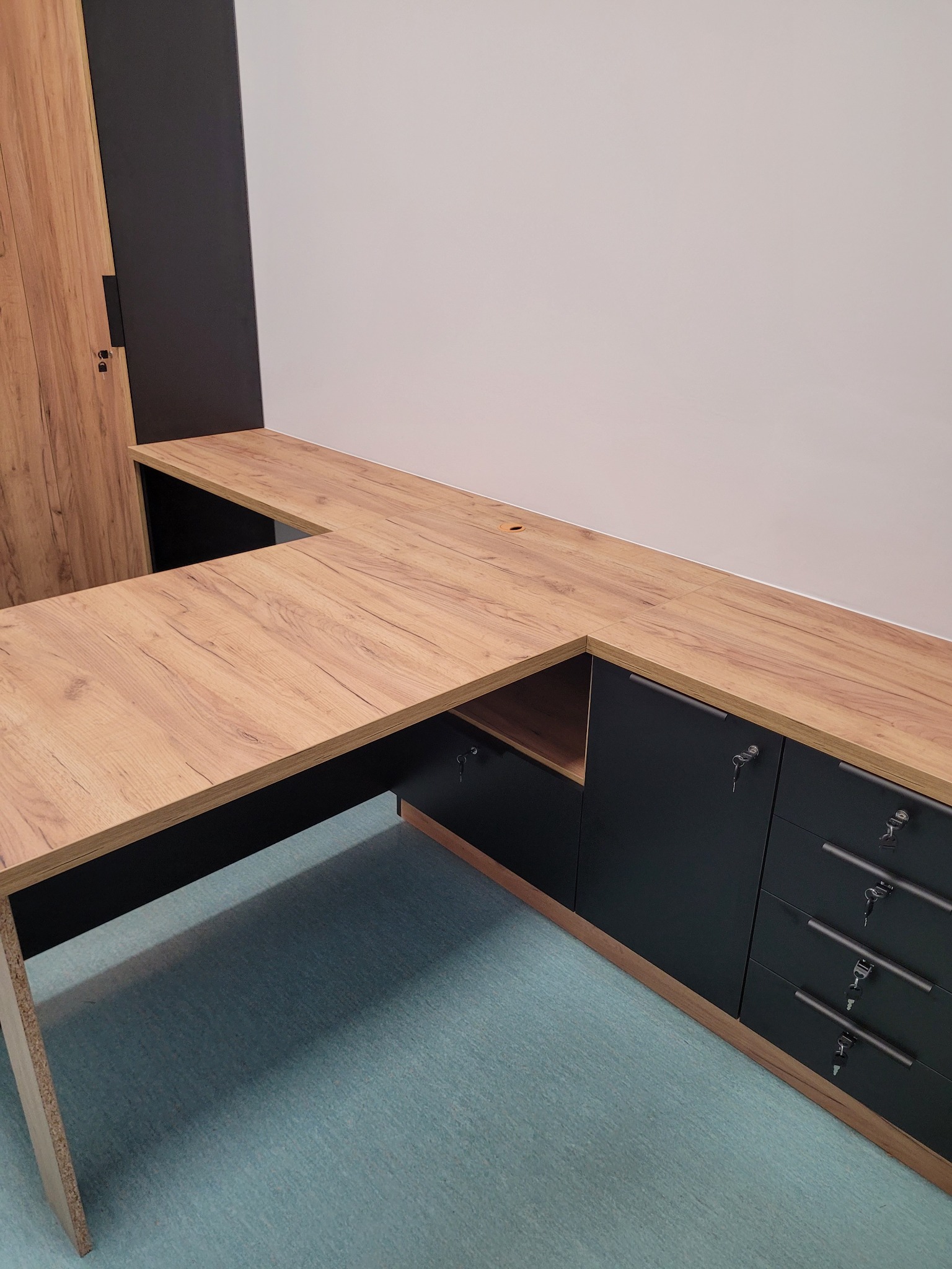 Kątowe biurko z blatem imitującym drewno i czarnymi szafkami z szufladami zamykanymi na klucz, widok z dołu.