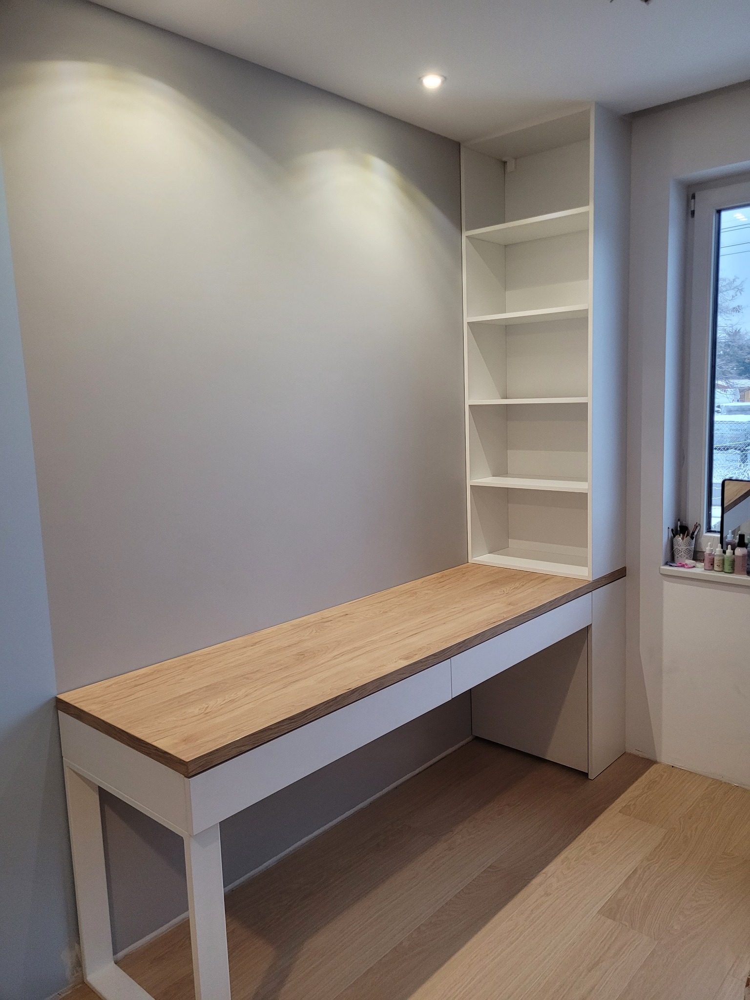 Biurko z drewnianym blatem zintegrowane z białą szafką z półkami, ustawione przy ścianie i oświetlone punktowym światłem.