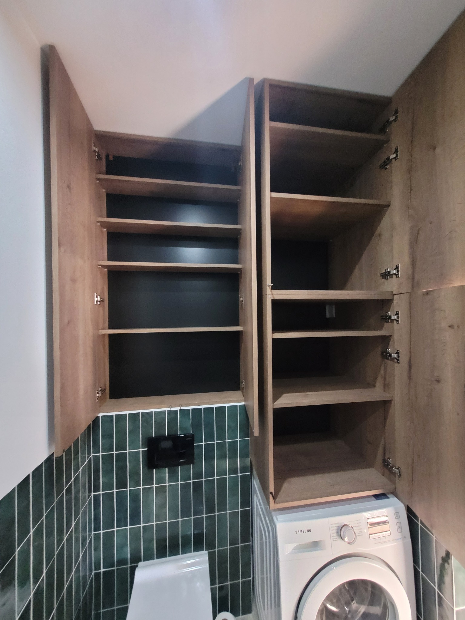 Drewniane szafki łazienkowe z półkami, zamontowane nad pralką i toaletą, na tle ściany wyłożonej ciemnozielonymi płytkami.