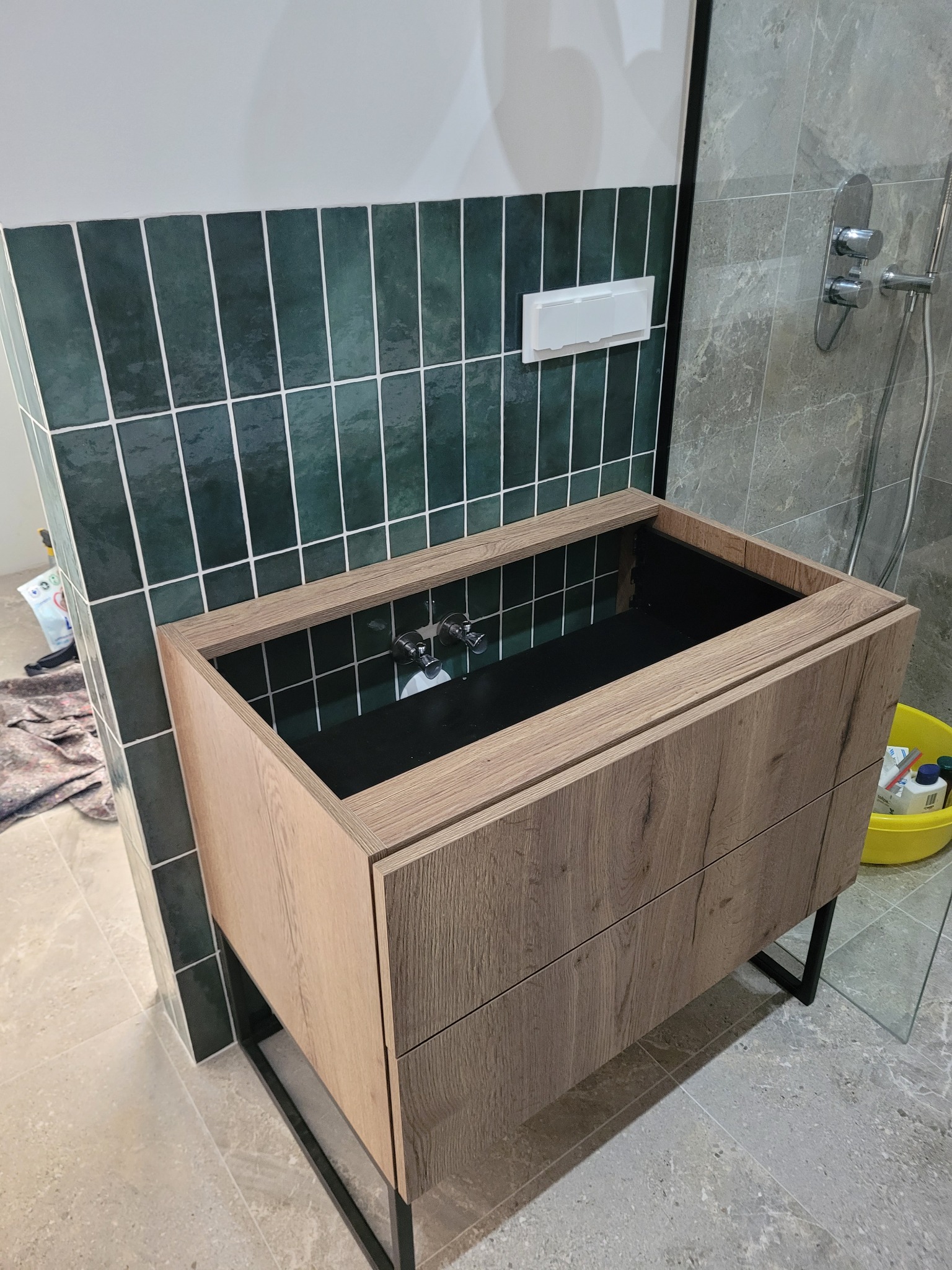 Łazienkowa szafka pod umywalkę z drewnianym frontem i czarnym wnętrzem, na tle zielonych i szarych płytek w nowoczesnej łazience.