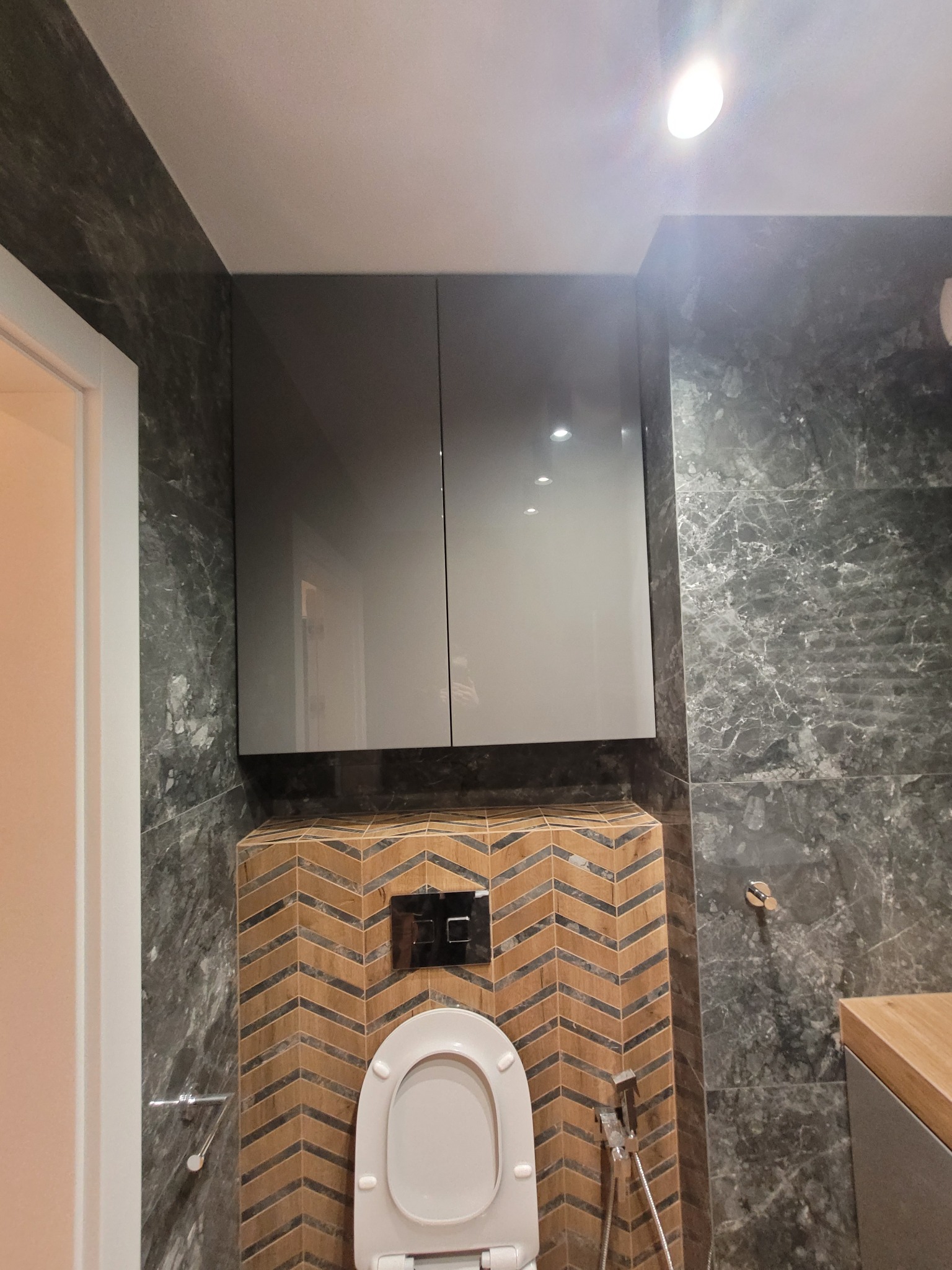 Nowoczesna toaleta podwieszana z szarymi szafkami nad nią, wykończona płytkami imitującymi marmur i drewnem ułożonym w jodełkę.