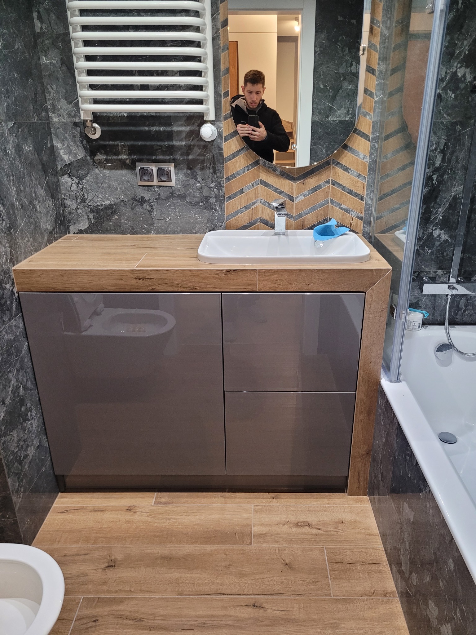 Nowoczesna łazienka z szafką pod umywalką w kolorze taupe, drewnianym blatem i lustrem z wzorem w jodełkę. W odbiciu lustra widoczna osoba robiąca zdjęcie. Na ścianie grzejnik drabinkowy...