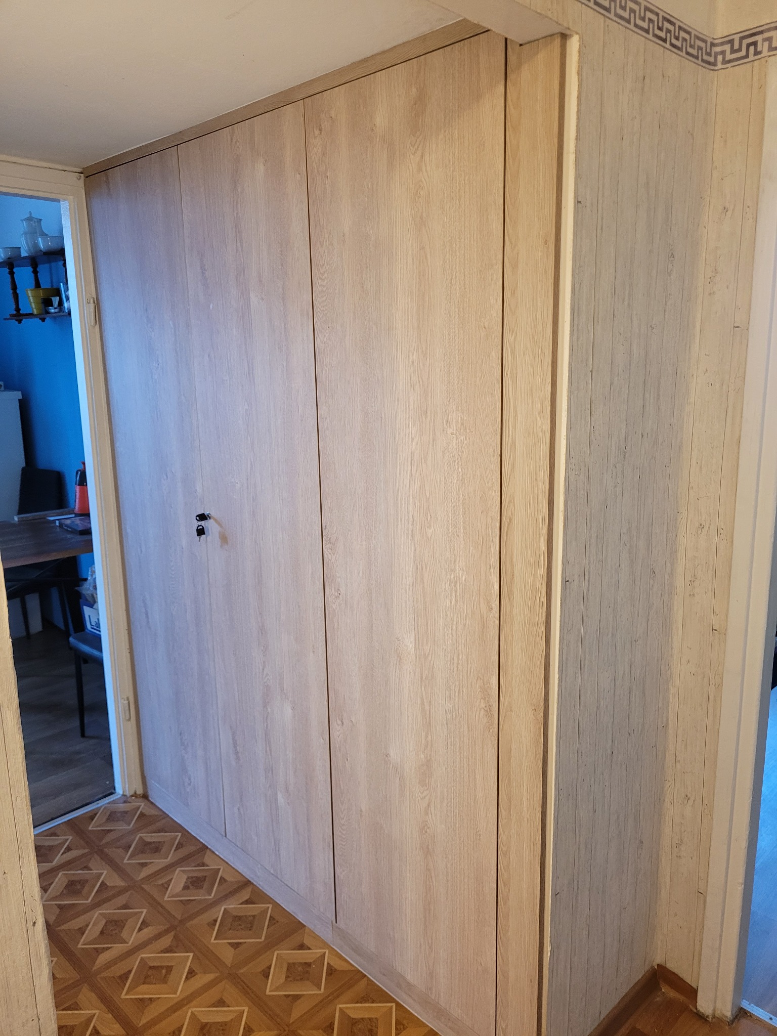 Jasna, drewnopodobna szafa wnękowa z zamkiem na klucz, przylegająca do ściany z tapetą w delikatne pionowe paski i ozdobnym greckim fryzem pod sufitem, widoczna z perspektywy korytarza z wzorzystą...