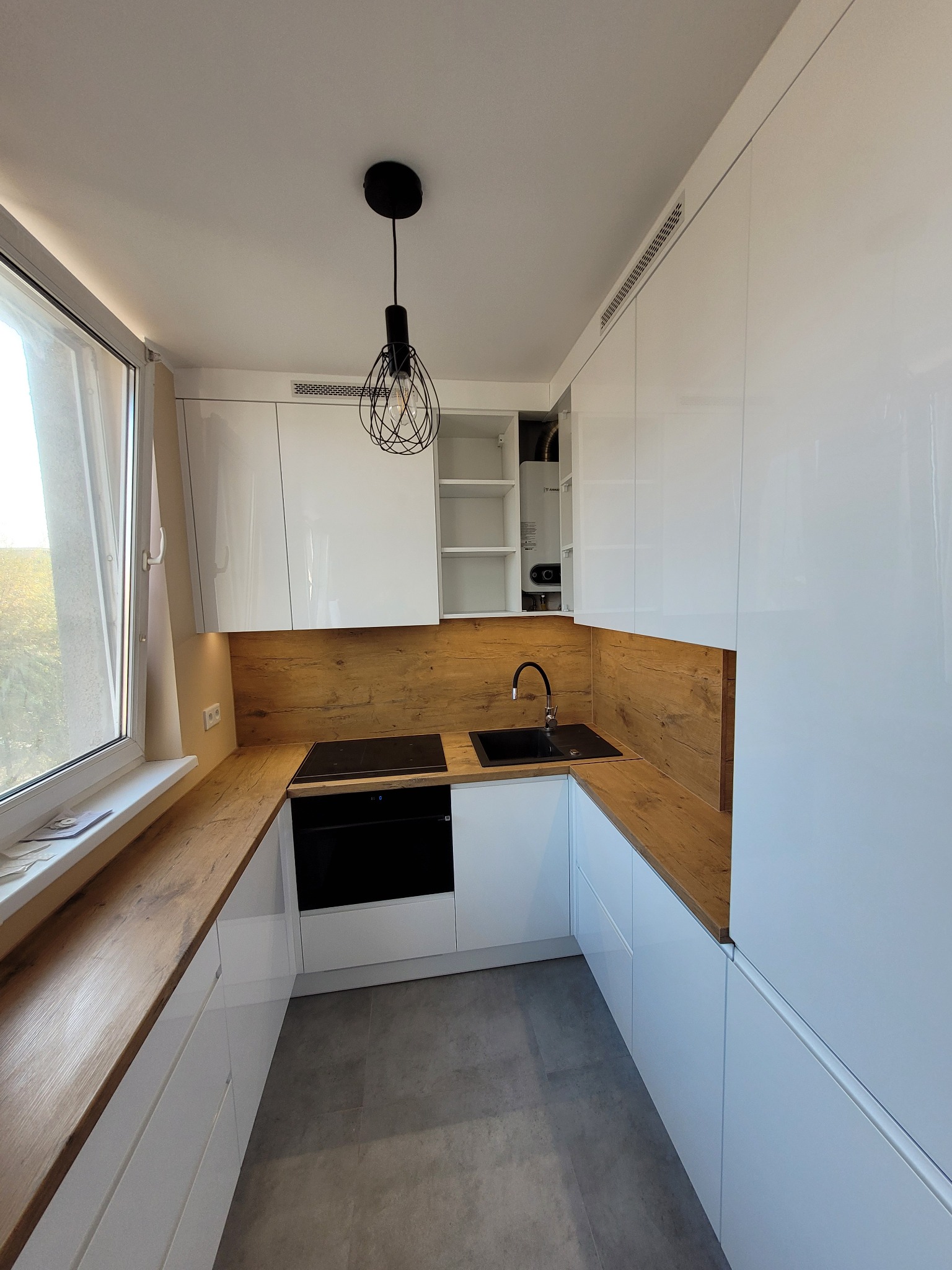 Nowoczesna kuchnia z białymi, błyszczącymi szafkami, drewnianym blatem i panelem ściennym, czarnym zlewem i płytą indukcyjną, oświetlona dekoracyjną lampą wiszącą.