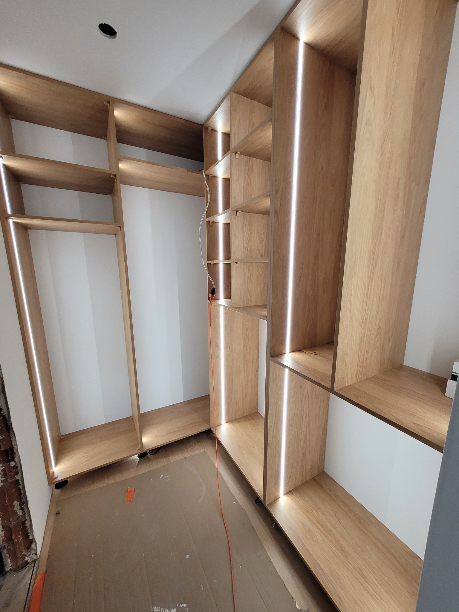 Wnętrze garderoby w trakcie montażu, z drewnianymi półkami i wbudowanym oświetleniem LED, widoczne kable i zabezpieczenia podłogi.
