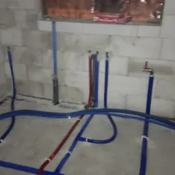 Surowy stan łazienki w trakcie prac instalacyjnych: widoczne rury wodne w kolorach niebieskim i czerwonym, prowadzone po podłodze i ścianach z betonu komórkowego. Na ścianie zamontowany zawór.
