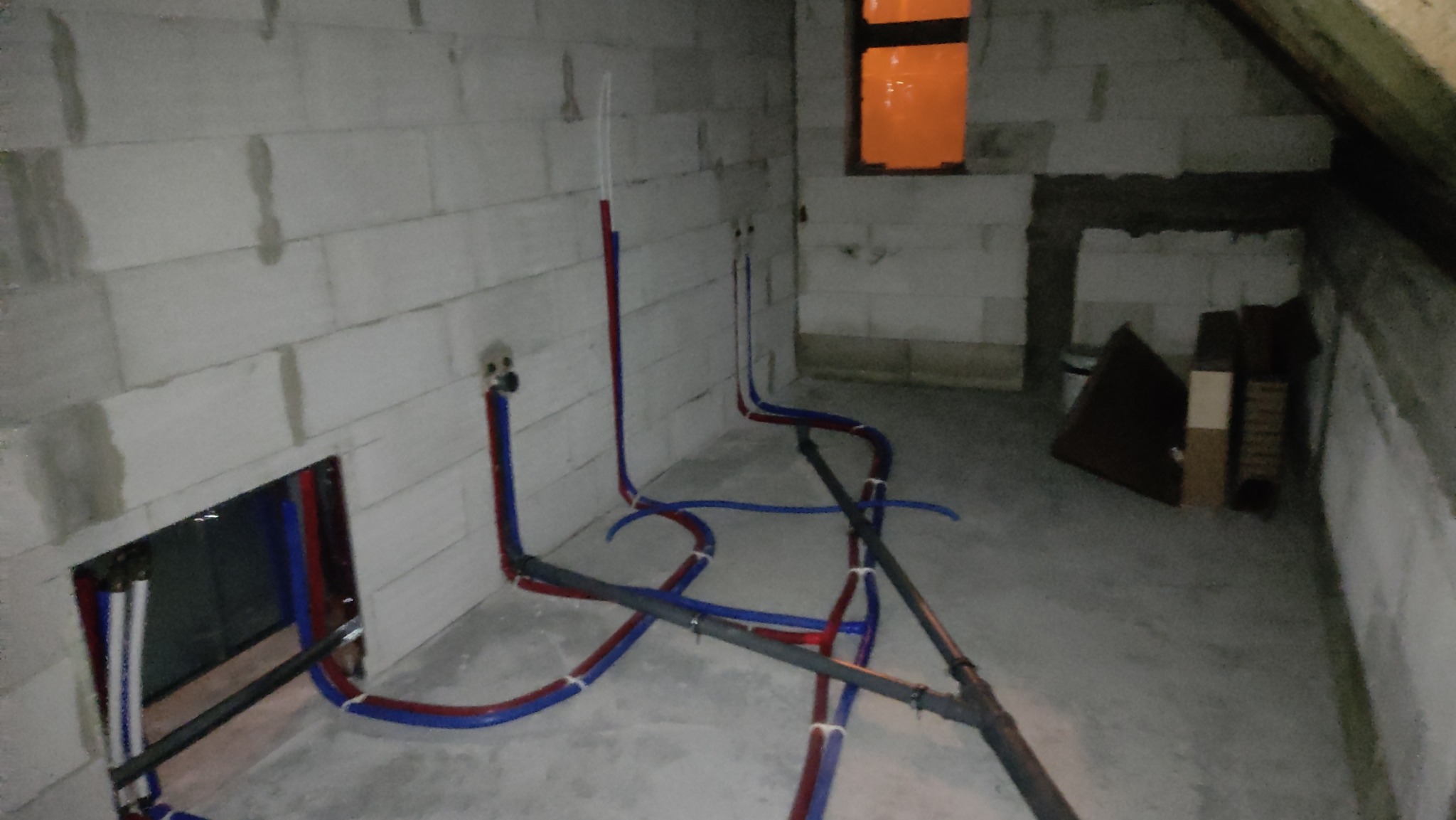 Surowy stan poddasza z widocznymi, świeżo rozprowadzonymi rurami instalacji wodnej i kanalizacyjnej, częściowo ukrytymi w bruzdach ściennych, z oknem w tle.