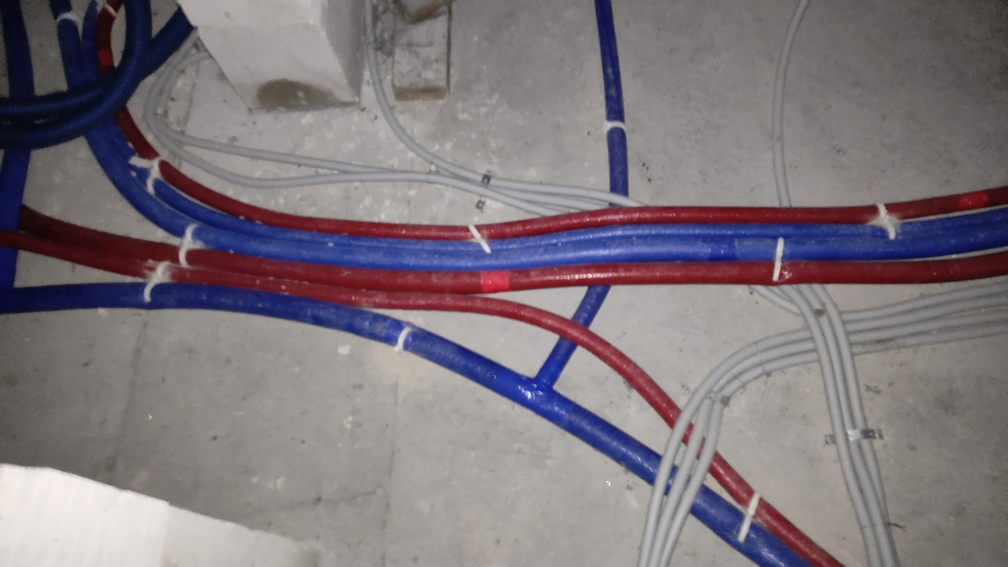 Wiązka rur instalacyjnych w kolorach czerwonym i niebieskim, połączonych białymi opaskami, biegnących po szarej, betonowej posadzce w surowym stanie.