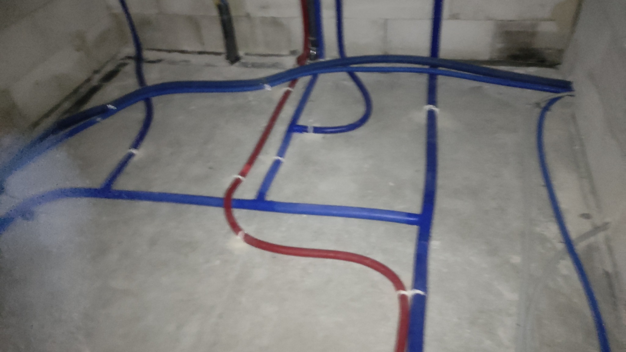 Wnętrze w trakcie prac instalacyjnych: niebieskie i czerwone rury prowadzone po szarej, betonowej podłodze, przymocowane białymi opaskami, ściany z białych bloczków.