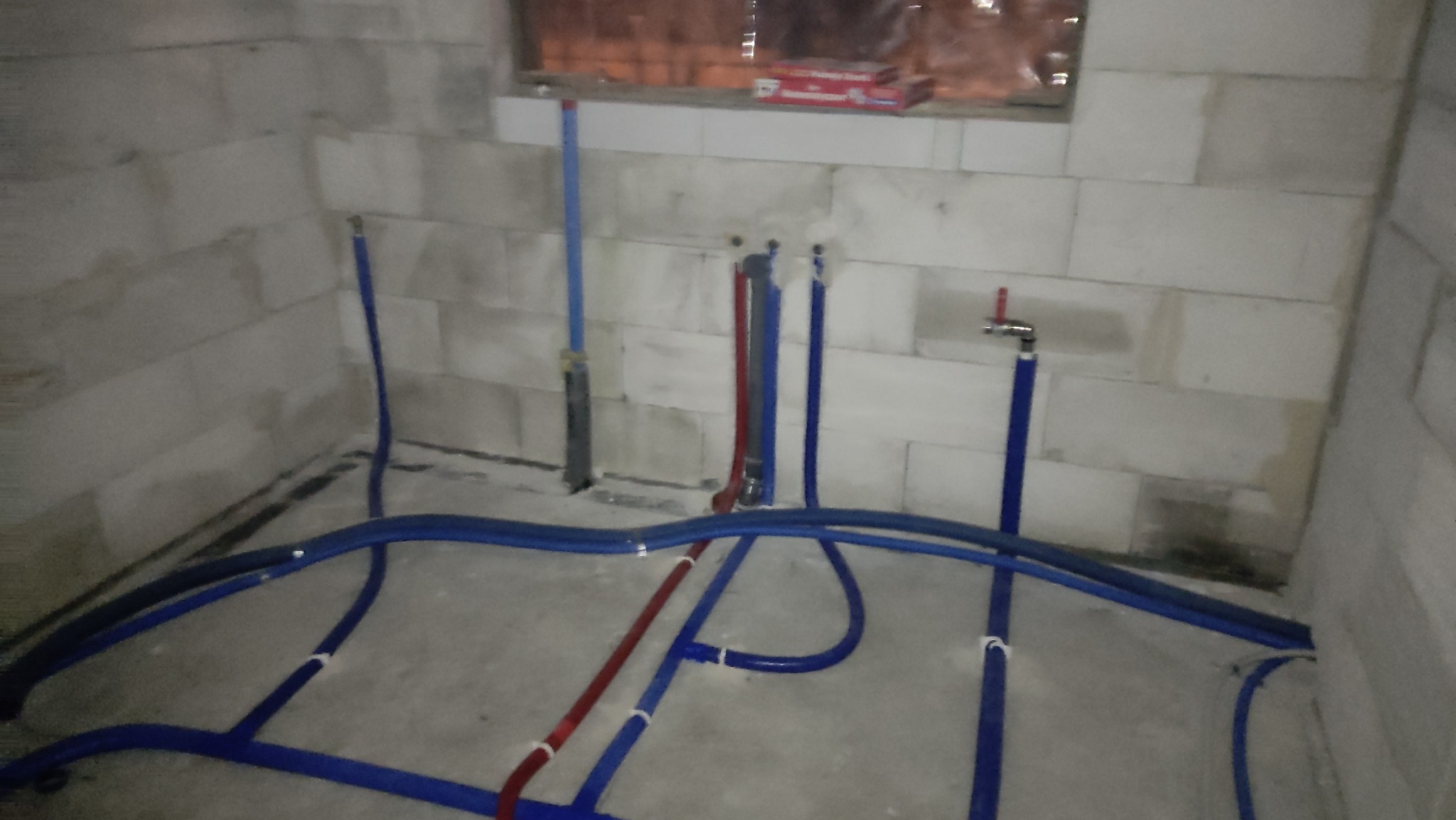Surowy stan łazienki w trakcie prac instalacyjnych: widoczne rury wodne w kolorach niebieskim i czerwonym, prowadzone po podłodze i ścianach z betonu komórkowego. Na ścianie zamontowany zawór.