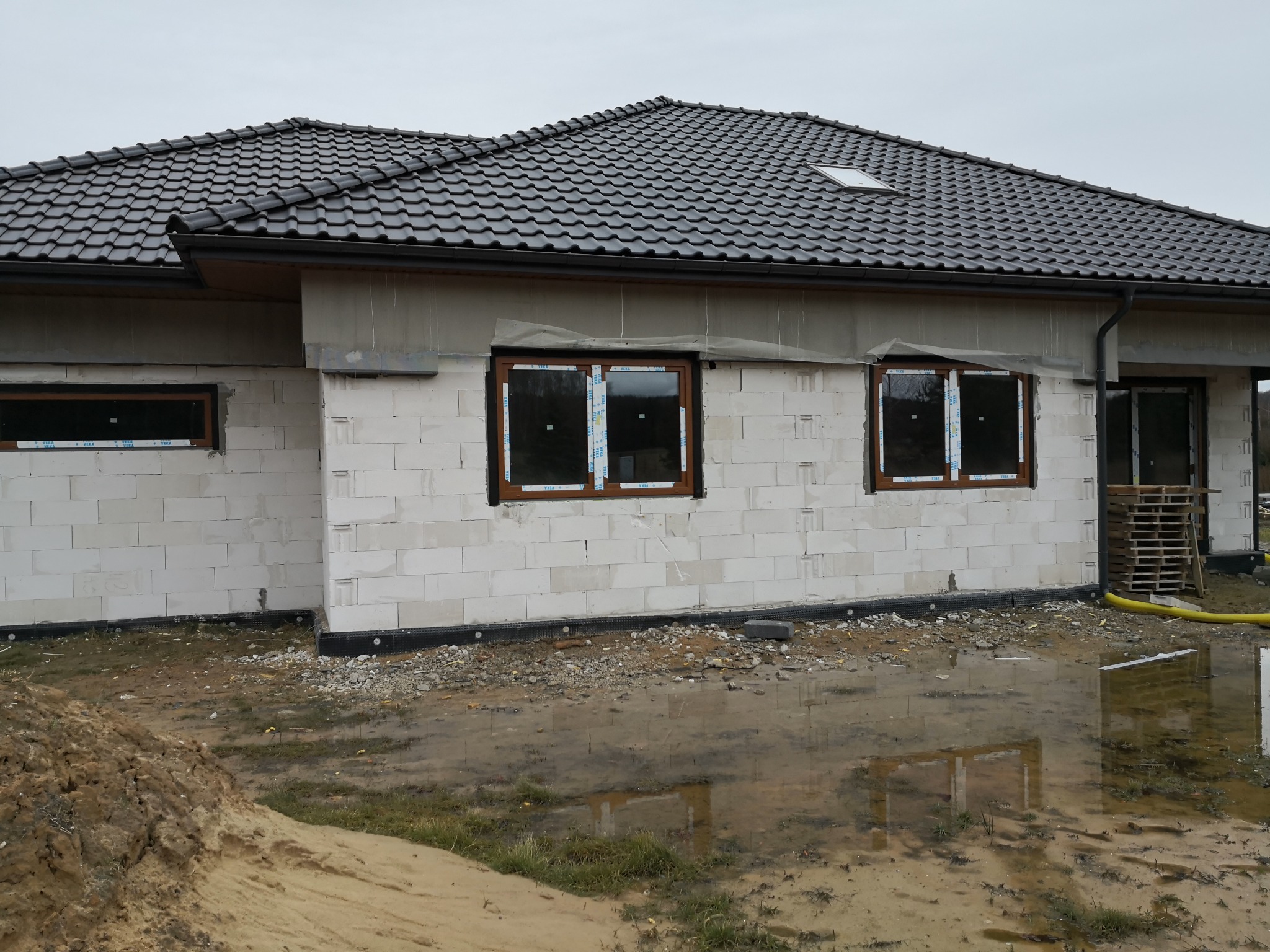 Surowy stan budynku w trakcie budowy, widoczne zamontowane okna z brązowymi ramami, ściany z białych bloczków, czarny dach z dachówki, ziemia po deszczu.